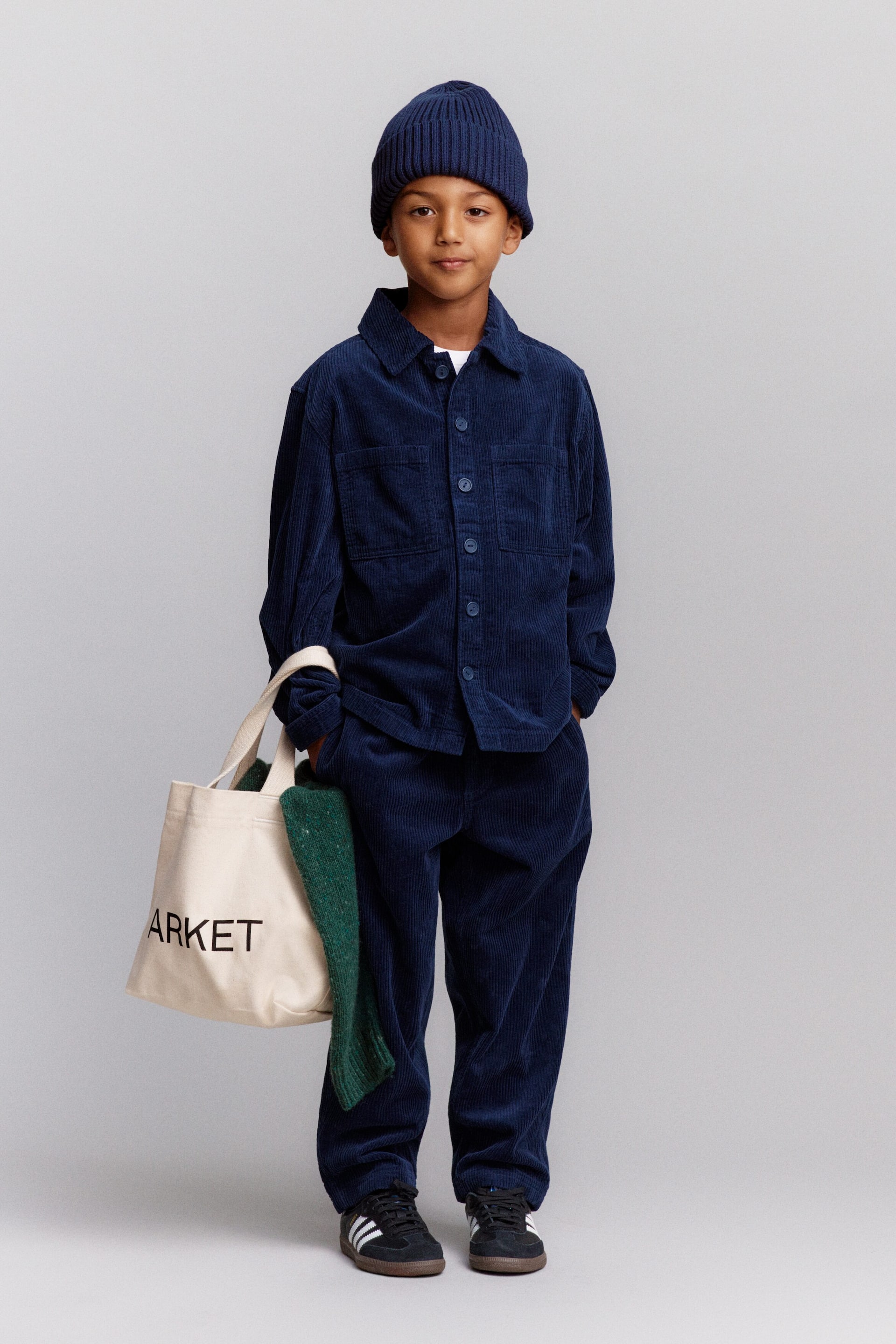 Grösseres Bild ansehen: Ein Kind steht frontal, trägt ein farblich abgestimmtes marineblaues Cord-Overshirt mit breiter Rippe und eine locker geschnittene Hose, kombiniert mit einer gerippten Baumwoll-Beanie. Es hält eine cremefarbene Tote-Bag aus Baumwoll-Canvas mit gedrucktem ARKET-Logo, an deren Seite ein grün gesprenkelter Wollpullover drapiert ist.