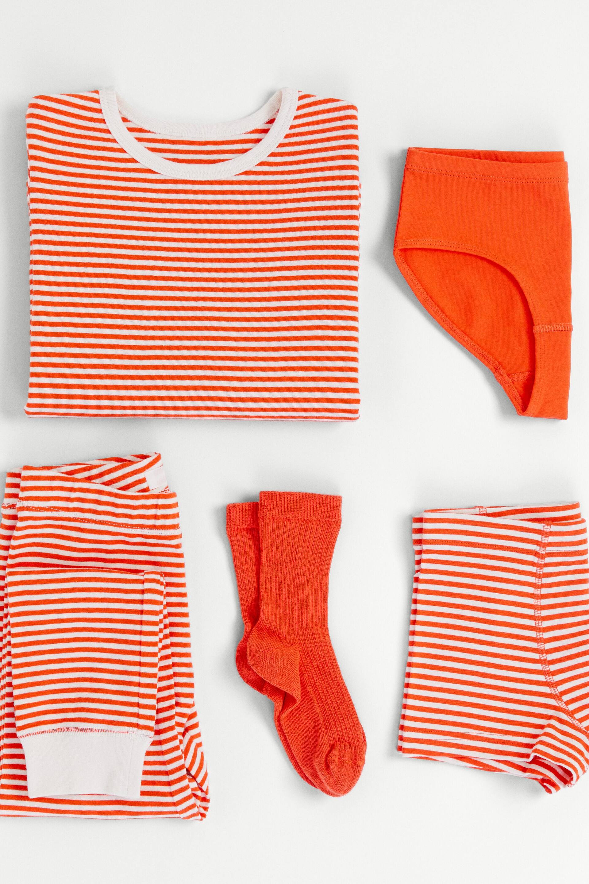 Vis større bilde: En brettet topp med oransje og hvite striper og hvit rund hals vises sammen med ensfargede oransje briefs. Et par brettede bukser med oransje og hvite striper og hvite ribbestrikkede mansjetter presenteres ved siden av ensfargede oransje ribbestrikkede sokker og brettede shorts med oransje og hvite striper.