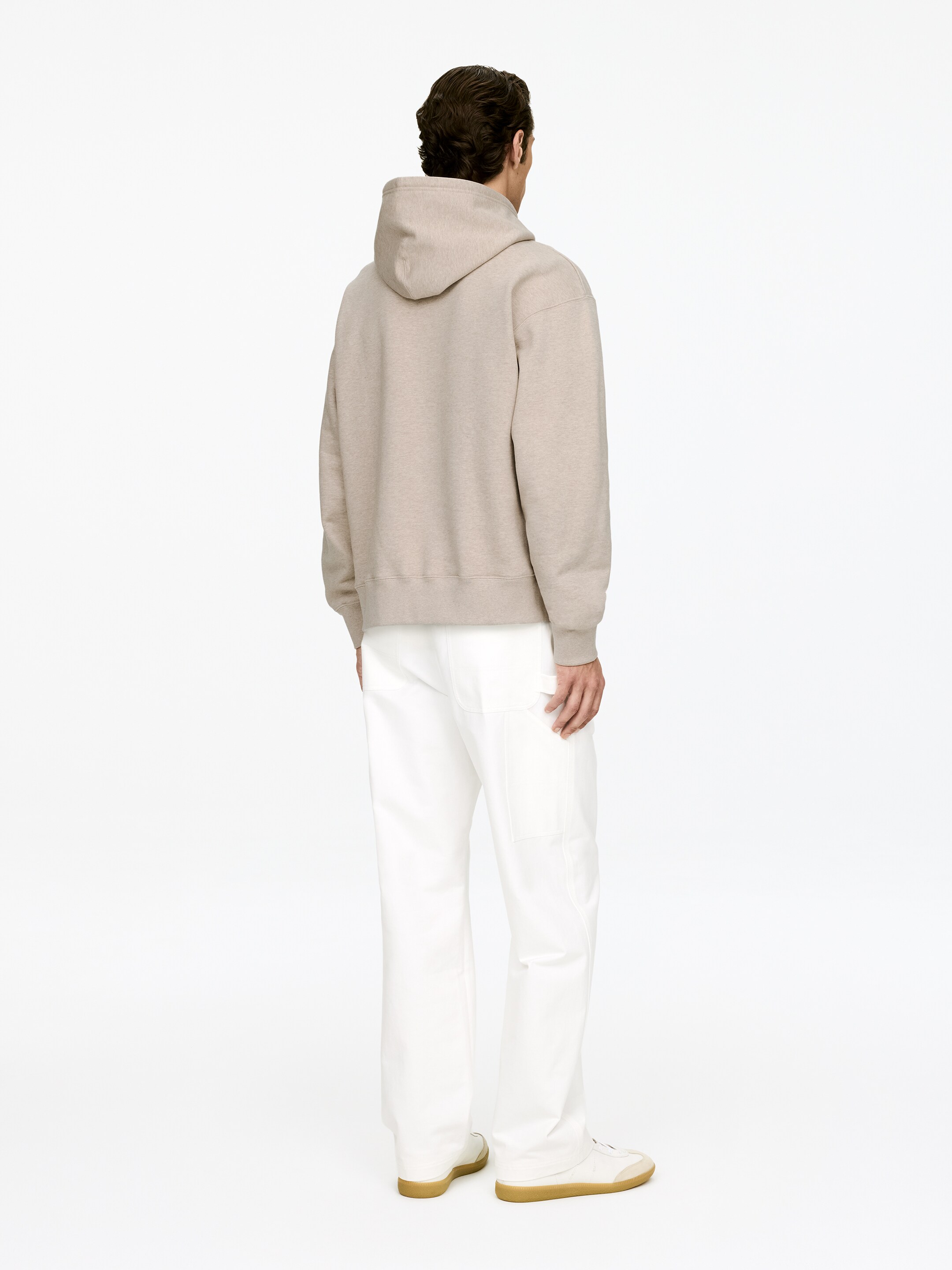 Visualizza immagine più grande: Una felpa con cappuccio beige chiaro melangiato, caratterizzata da una vestibilità comoda, un cappuccio, polsini a coste e un orlo a coste, viene mostrata di spalle, indossata sopra pantaloni bianchi a gamba dritta con dettagli di cucitura visibili e sneakers bianche con suole color cuoio.