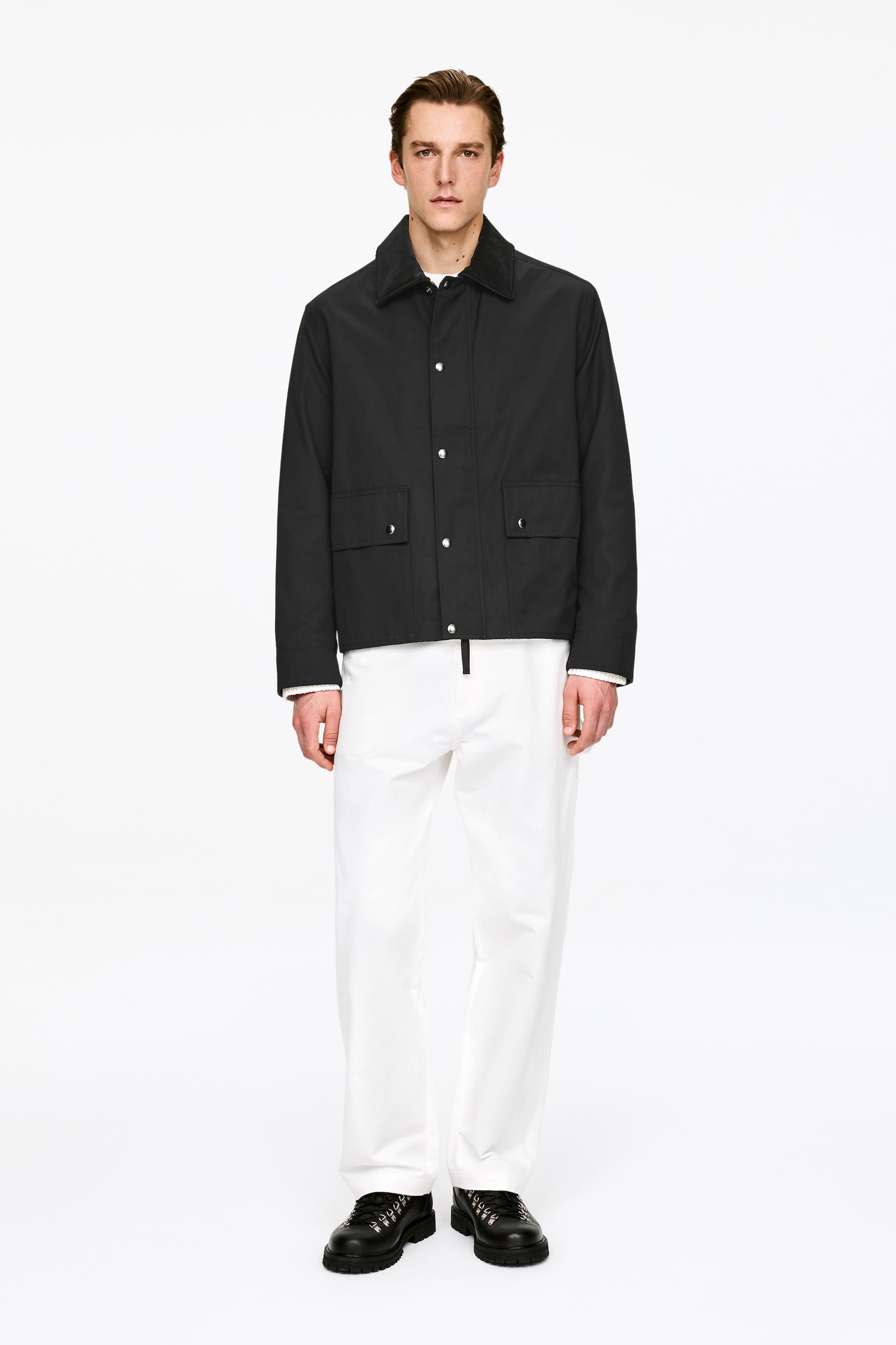 View larger image: The black jacket is shown front-facing, featuring a dark contrasting collar and a front closure with silver-toned snap fastenings. It also includes two rectangular flap pockets with snap closures on the lower front and is styled over a white long-sleeved shirt.