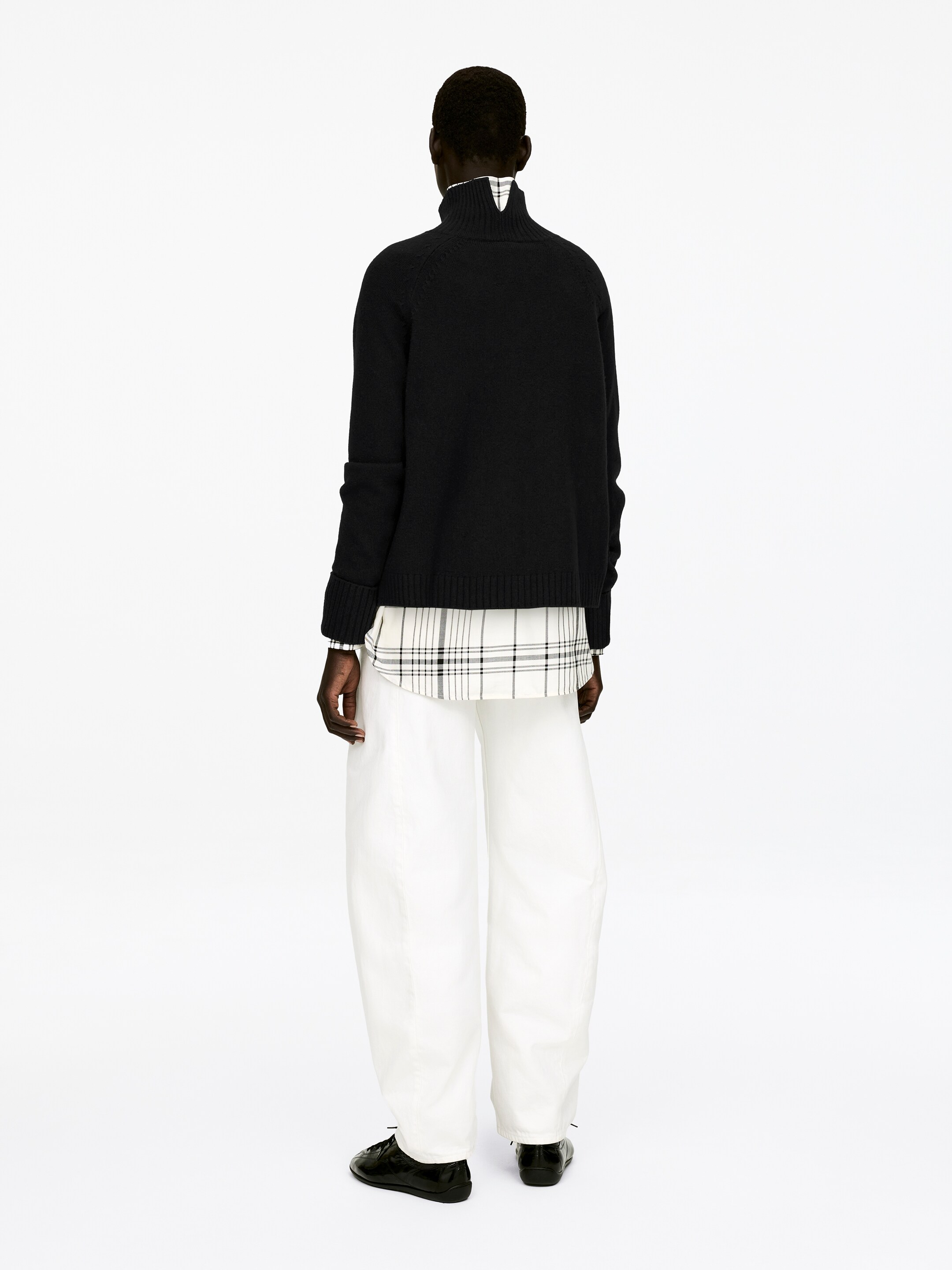 View larger image: A black roll-neck jumper, featuring raglan sleeves and folded cuffs, is being worn by the model. The jumper is layered over a plaid shirt, which is visible at the collar and hem.