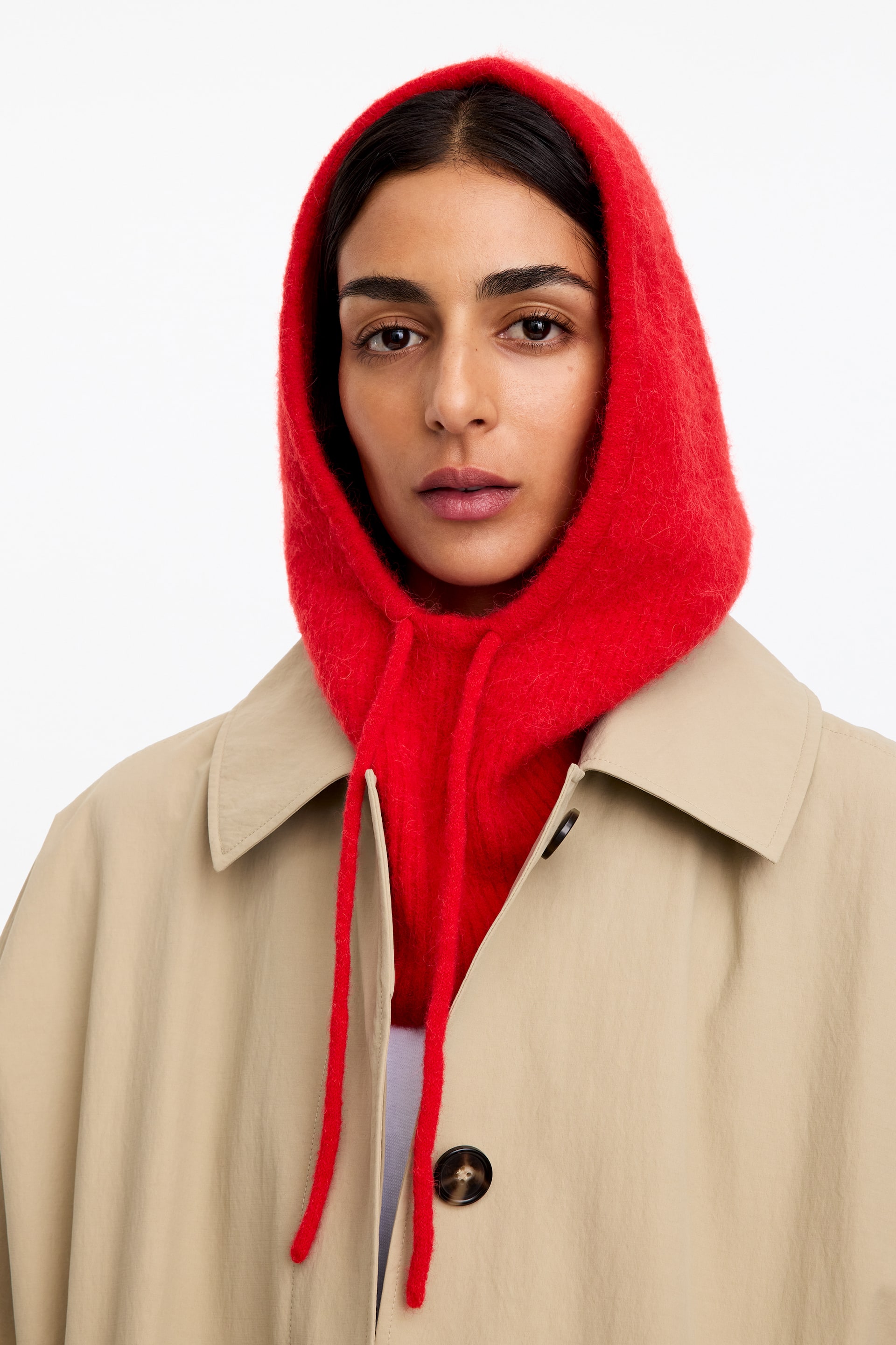 Größeres Bild anzeigen: Das Model trägt eine rote gestrickte Kapuze mit langen Kordeln, die unter einem beigen Trenchcoat mit klassischem Kragen getragen wird.