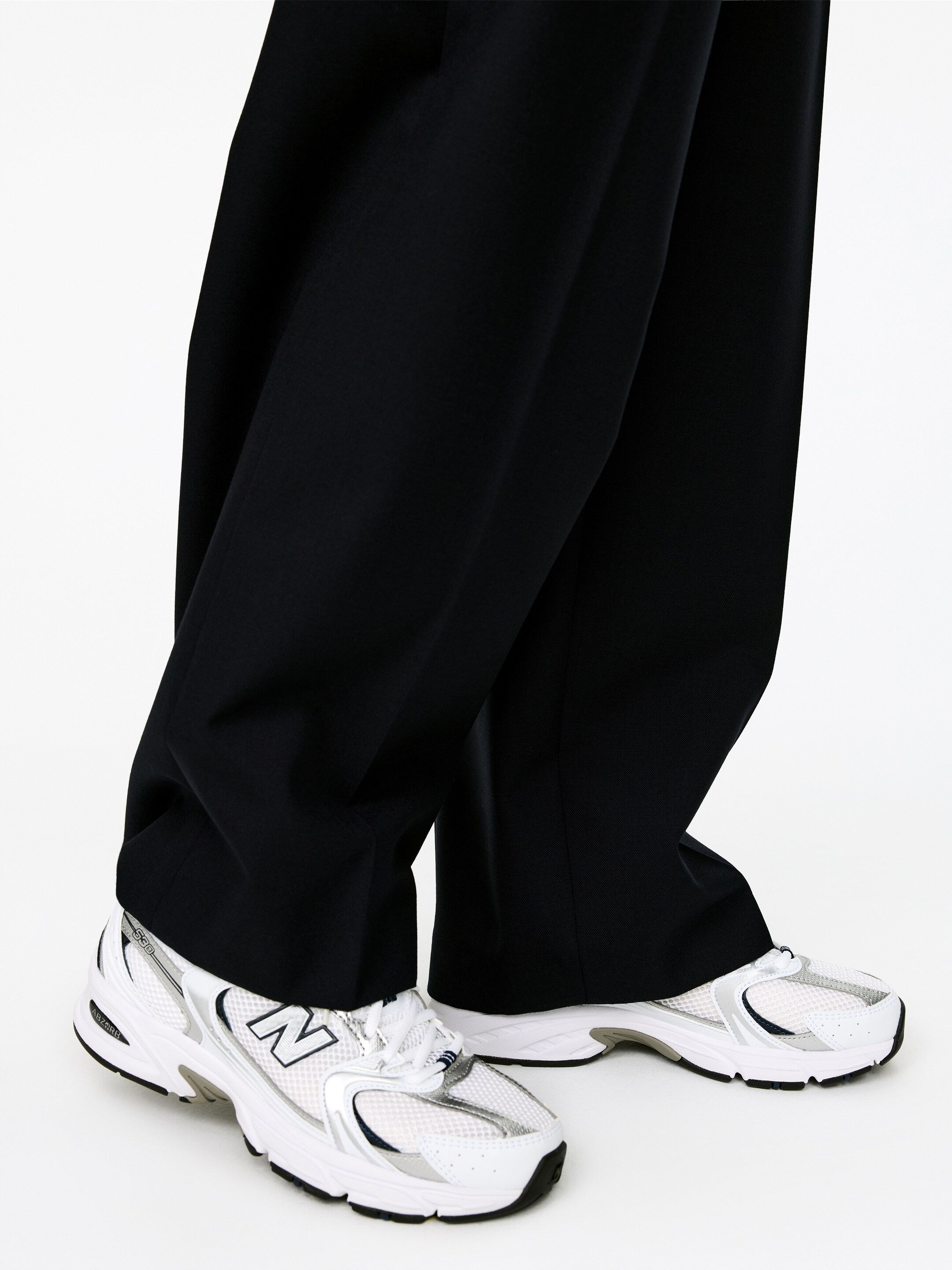 Ver imagen más grande: Pantalones negros de pierna ancha caen sobre unas zapatillas New Balance 530 blancas. Las zapatillas presentan partes superiores de malla blanca, superposiciones metálicas plateadas y suelas negras, con los pantalones cubriendo parcialmente sus secciones superiores.