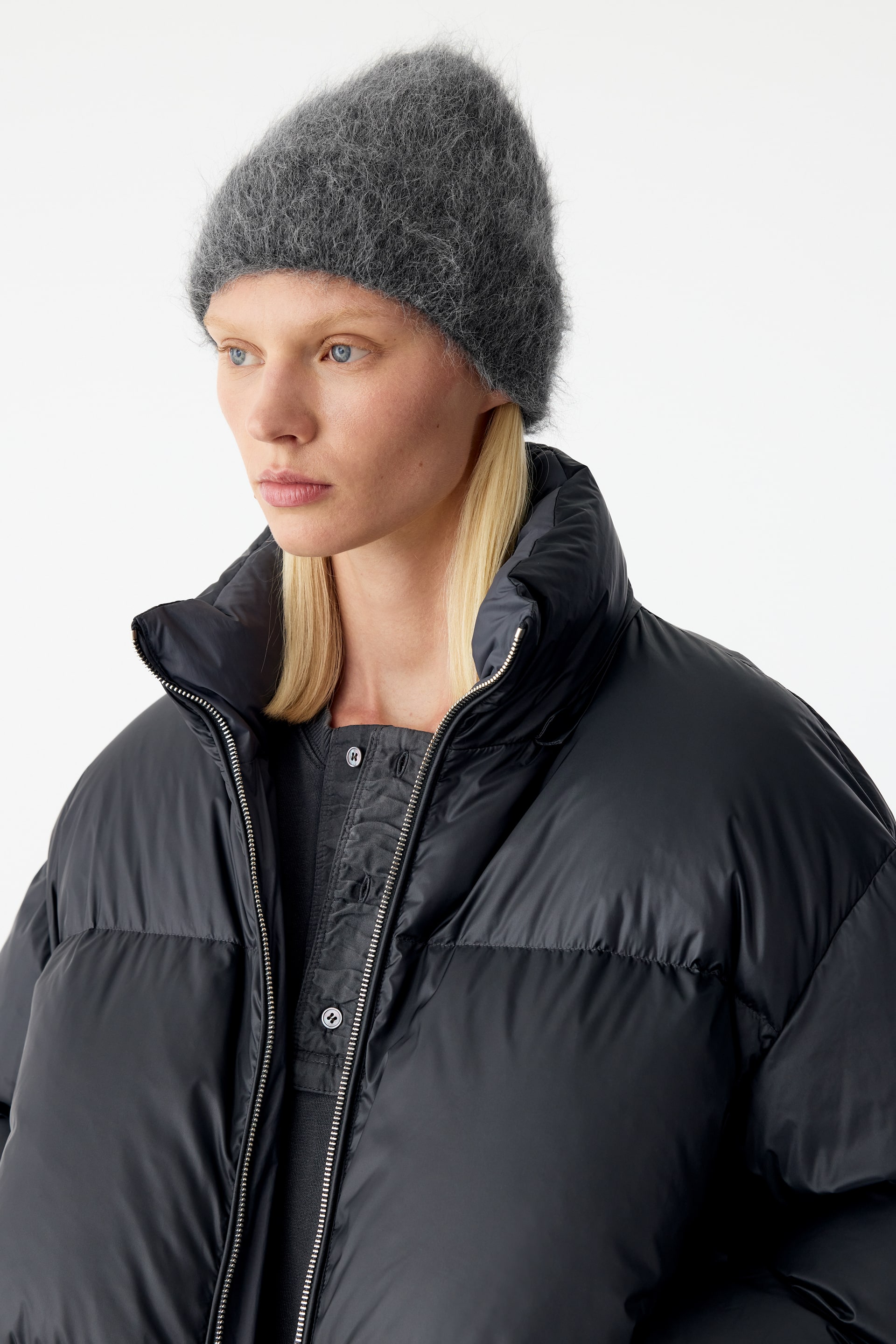 View larger image: A dark grey, fluffy beanie with a brushed knitted texture is being worn by the model. The beanie is styled with a high-collared black puffer jacket.
