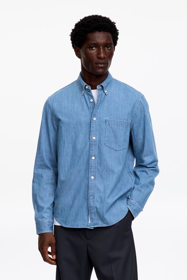 Viene presentata una camicia di denim azzurro, vista frontalmente, con colletto button-down, abbottonatura completa con bottoni bianchi e una tasca applicata sul petto a sinistra. È indossata con maniche lunghe che arrivano ai polsini abbottonati, sopra una T-shirt bianca. - 1