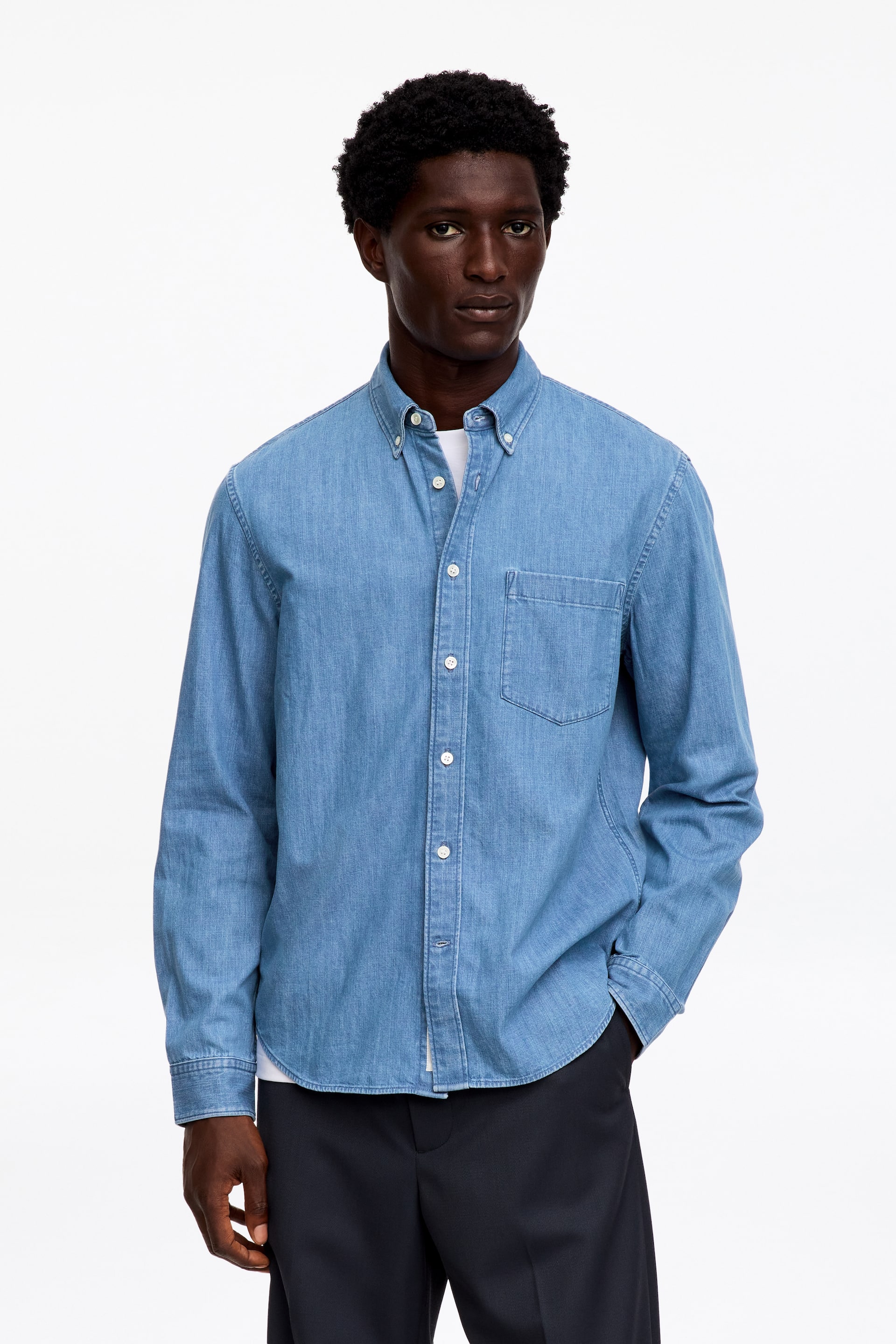 Größeres Bild anzeigen: Ein hellblaues Denim-Hemd wird präsentiert, frontal ausgerichtet, mit Button-down-Kragen, durchgehender Knopfleiste mit weißen Knöpfen und einer aufgesetzten Brusttasche auf der linken Brustseite. Es wird mit langen Ärmeln gezeigt, die bis zu den geknöpften Manschetten reichen, und über einem weißen T-Shirt getragen.