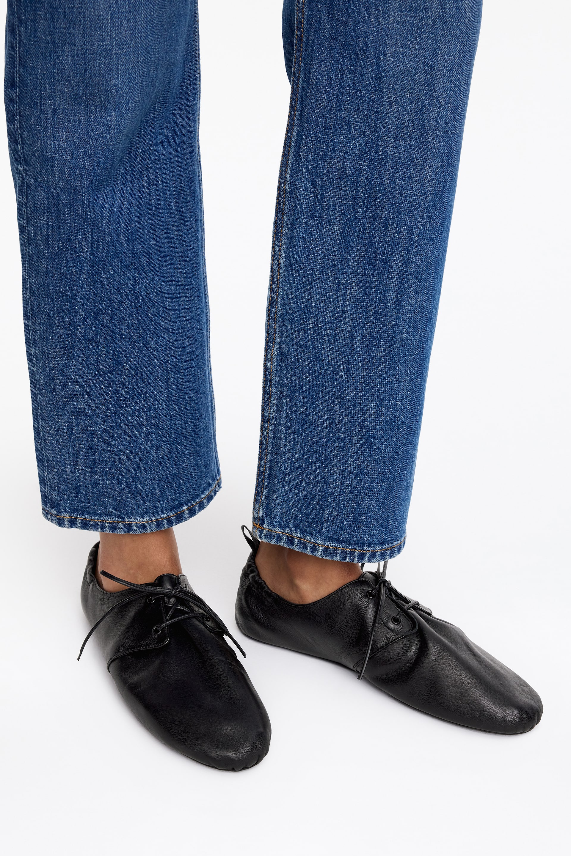 Visa större bild: Svarta läderskor presenteras från en något upphöjd framifrån-vinkel. Varje sko visar upp en mjuk, rynkad öppning runt ankeln och är säkrad med tunna svarta snören, som korsar över en slät vamp och leder till en mjukt rundad tå.