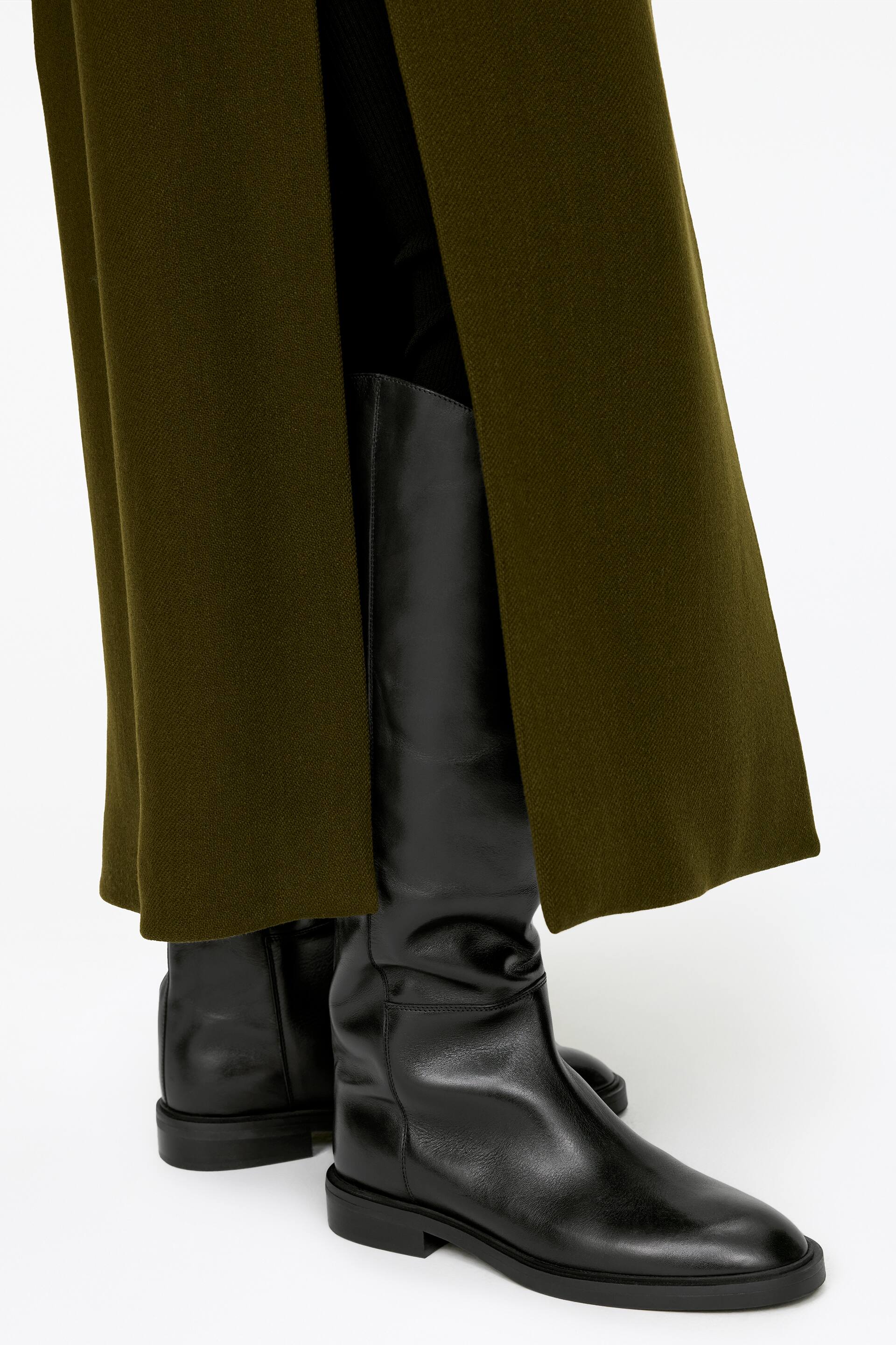 Größeres Bild ansehen: Schwarze, reitstiefel-inspirierte Stiefel werden vom Model getragen. Die Stiefel verfügen über ein glattes Lederobermaterial, eine abgerundete Spitze und einen niedrigen 2,5 cm hohen Absatz. Ein olivgrünes Kleidungsstück bedeckt teilweise die Stiefelschäfte.