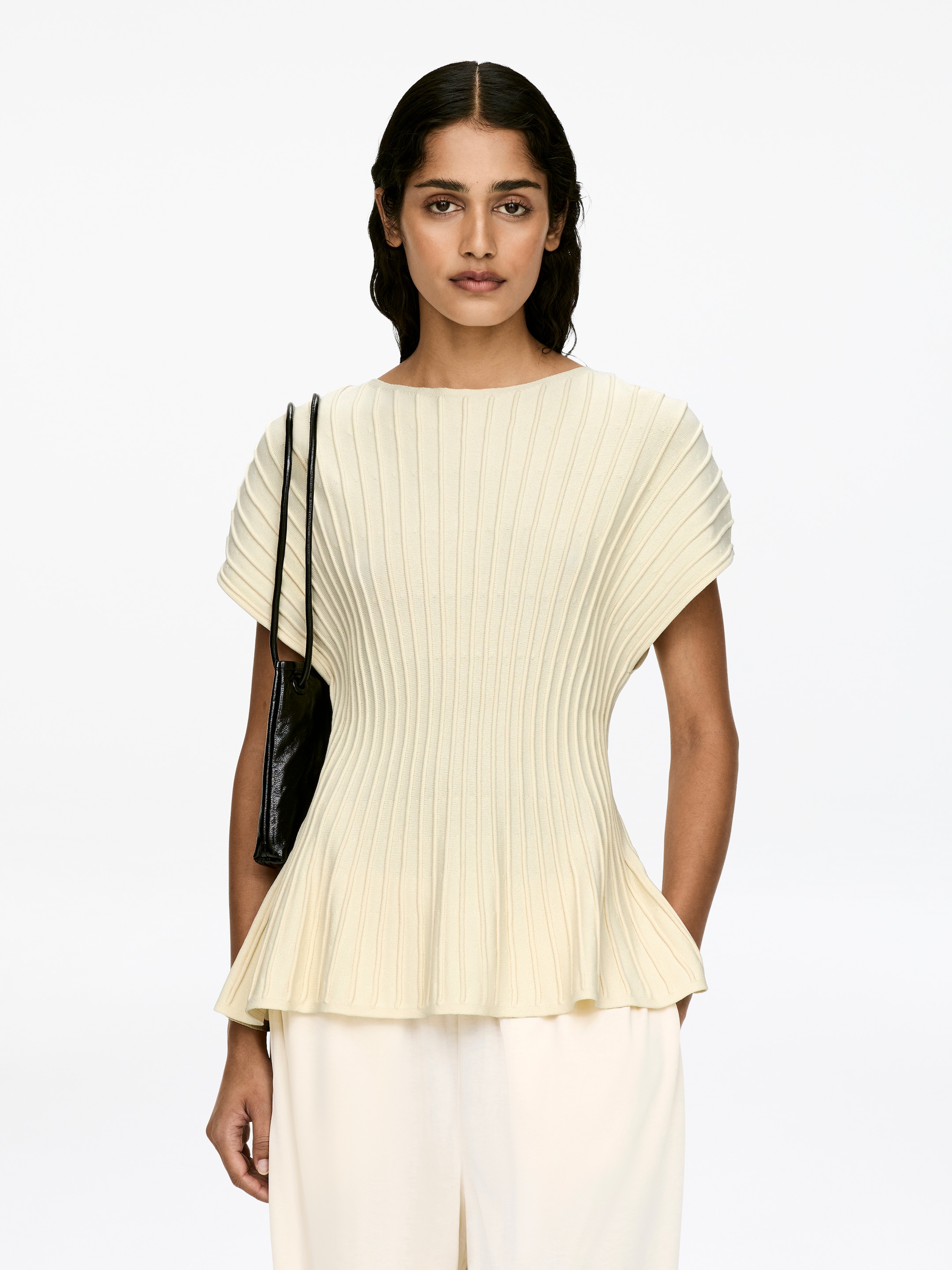 Visualizza immagine più grande: Un top a maglia fine di colore chiaro, caratterizzato da una cucitura orizzontale ottomana che crea una texture a coste e una silhouette a peplo, è indossato dalla modella. La modella indossa anche pantaloni di colore chiaro e porta una borsa a tracolla nera su una spalla.
