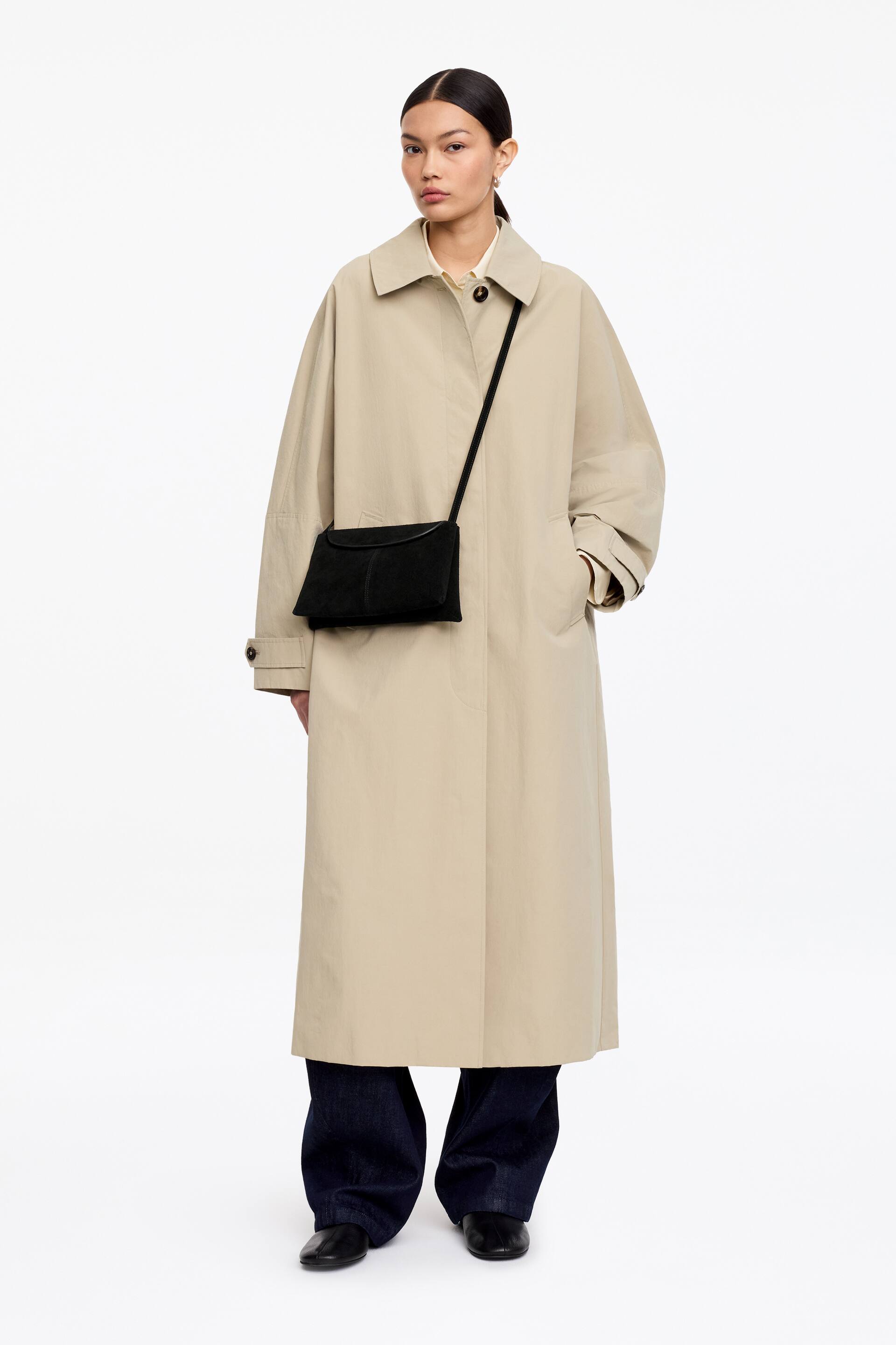 Visualizza immagine più grande: Un cappotto beige, lungo e ampio, è presentato di fronte, estendendosi fino alle caviglie. Presenta un colletto aperto, una chiusura centrale con un bottone scuro visibile, maniche larghe con polsini a linguetta con bottone e tasche laterali.