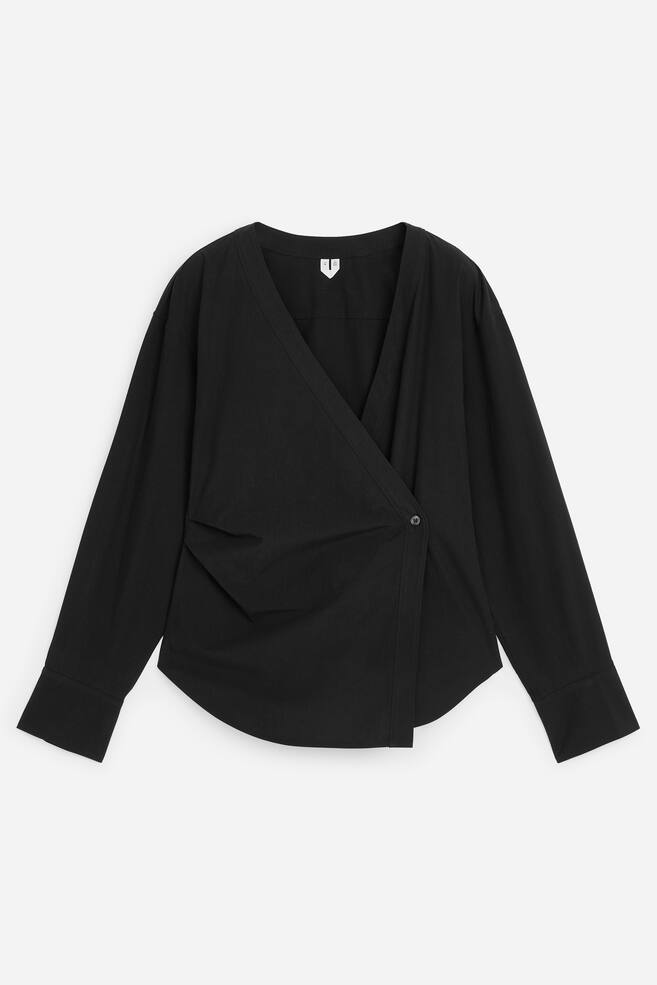Una blusa cruzada negra muestra su parte delantera, con un pronunciado escote en V formado por paneles frontales superpuestos que se aseguran con un único botón en el lado derecho del usuario. La blusa también presenta un pliegue recogido en el panel frontal izquierdo del usuario, junto con mangas largas con puños abotonados y un dobladillo curvado. - 1