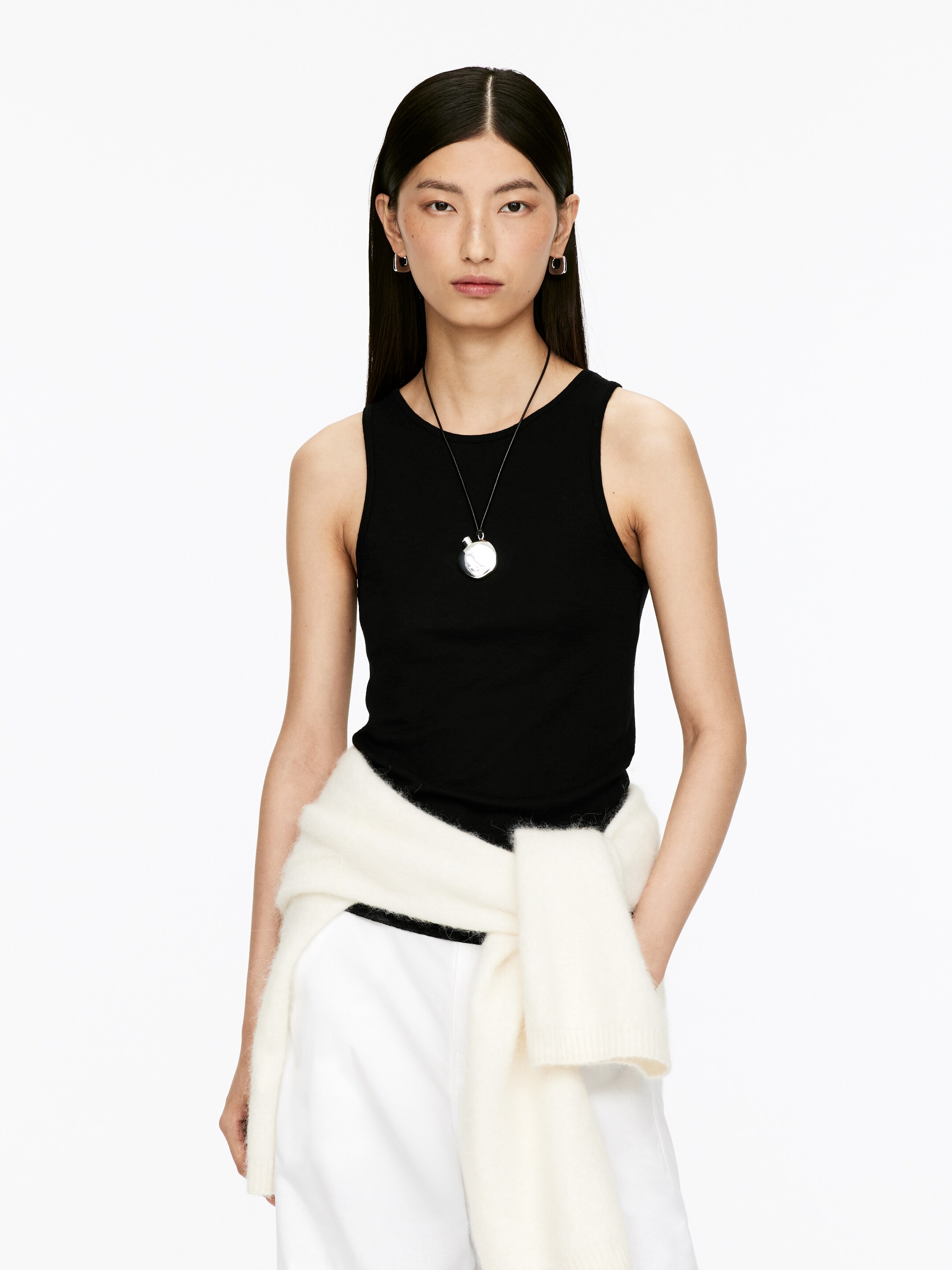 View larger image: A black fitted tank top is being worn, featuring a high crew neckline and a smooth knit texture. It is being styled with a silver-toned pendant resting on it and a cream-colored sweater tied around the waist over its lower portion.