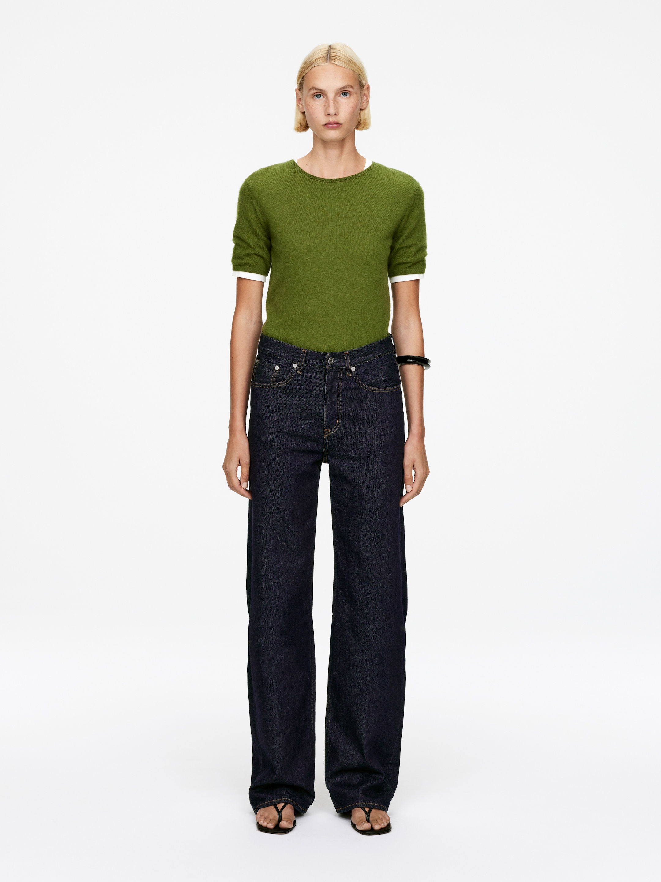 View larger image: An olive green, short-sleeved knit top is being worn, revealing a white layer at the crew neck and cuffs. It is being styled with dark wash, wide-leg jeans featuring contrast stitching, and black thong sandals.