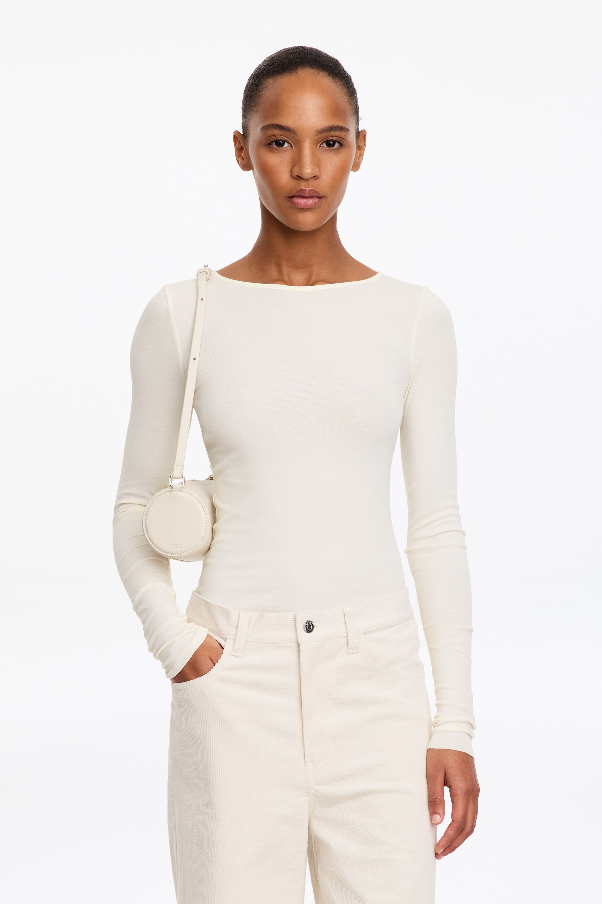 Visualizza l'immagine ingrandita: Un top a costine color crema, aderente, con ampia scollatura tonda e maniche lunghe, indossato dalla modella. Il top è infilato in pantaloni color crema coordinati ed è abbinato a una piccola borsa a tracolla rotonda.