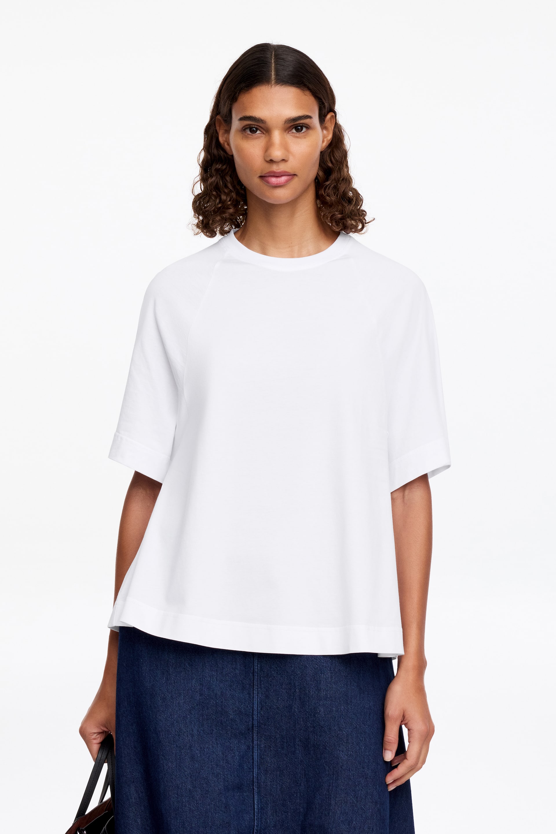Grösseres Bild ansehen: Ein weisses A-Linien-T-Shirt wird getragen, mit einem runden Halsausschnitt, Raglanärmeln und einer dezent ausgestellten Silhouette. Das Model trägt das T-Shirt mit einem dunkelblauen Rock und hält eine dunkle Tasche.