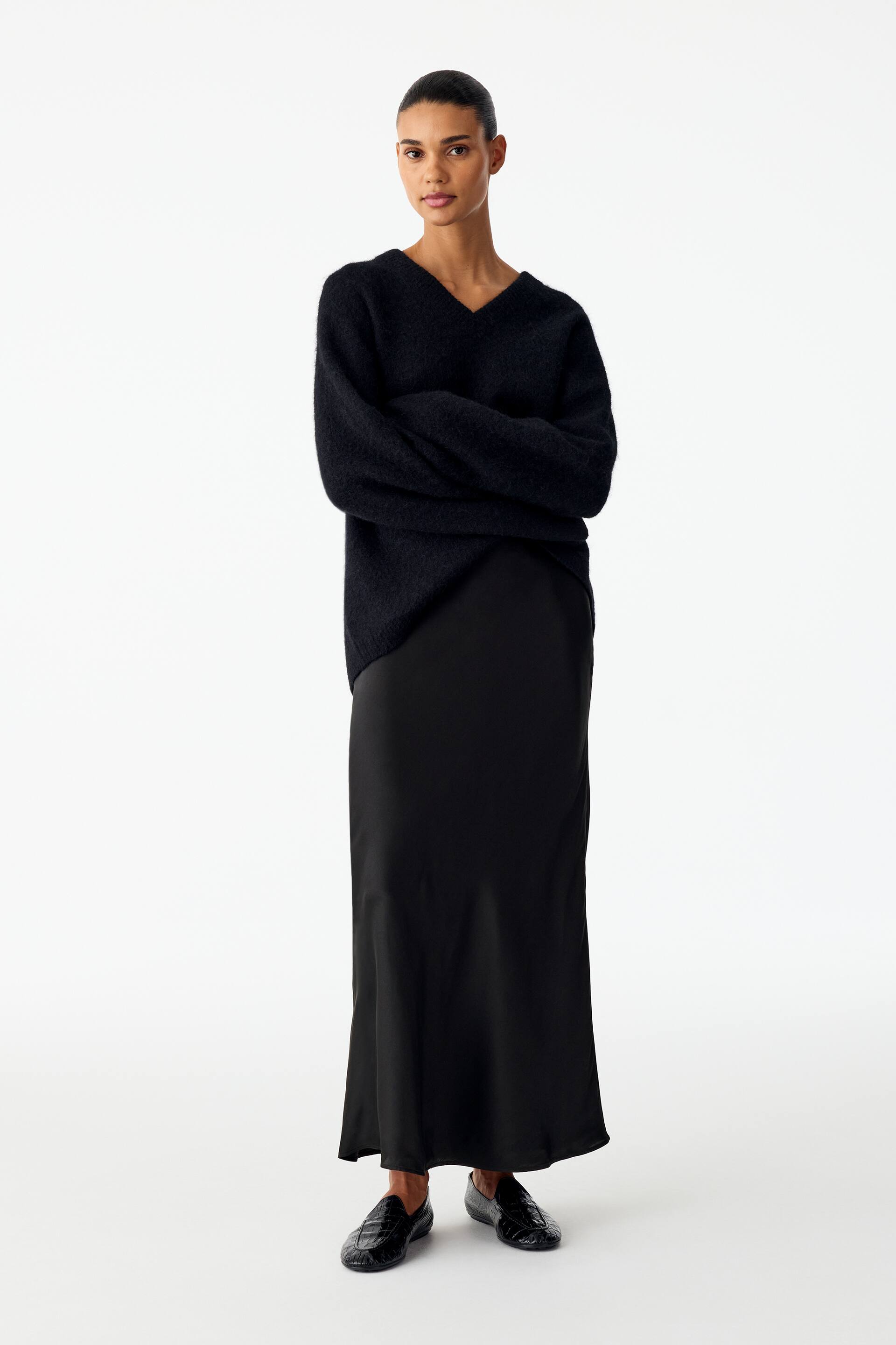 View larger image: A black silk satin maxi skirt is being worn by the model, draping fluidly to the ankles with a subtle sheen. The skirt is styled with a long-sleeved, black V-neck knitted jumper and black textured loafers.