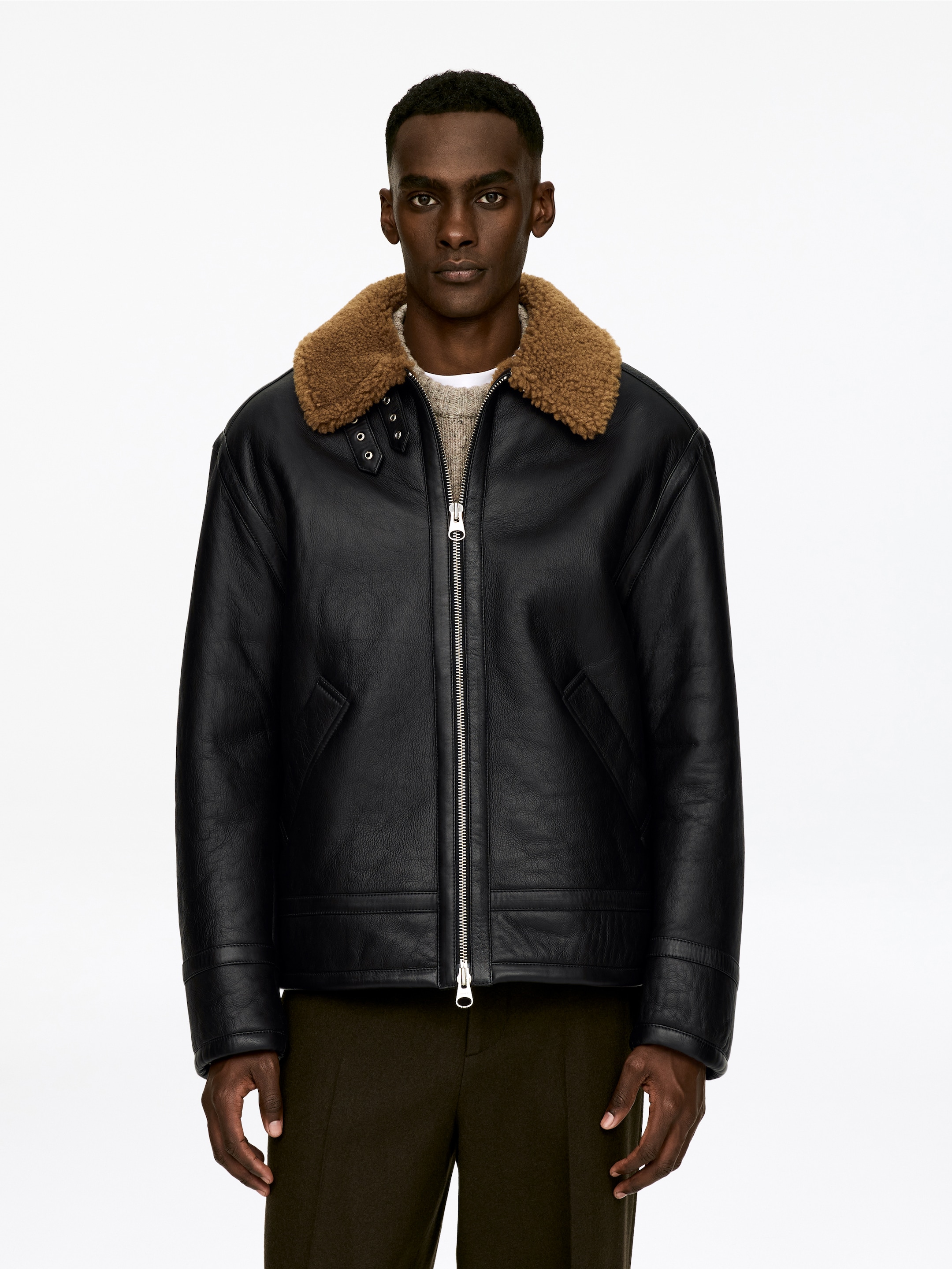 Agrandir l'image: Une veste en cuir noir est orientée vers l'avant, avec un col en shearling brun doté de détails à boucles et une fermeture éclair argentée le long du devant. Elle montre également deux poches en biais et est portée par-dessus un pull en tricot de couleur claire.