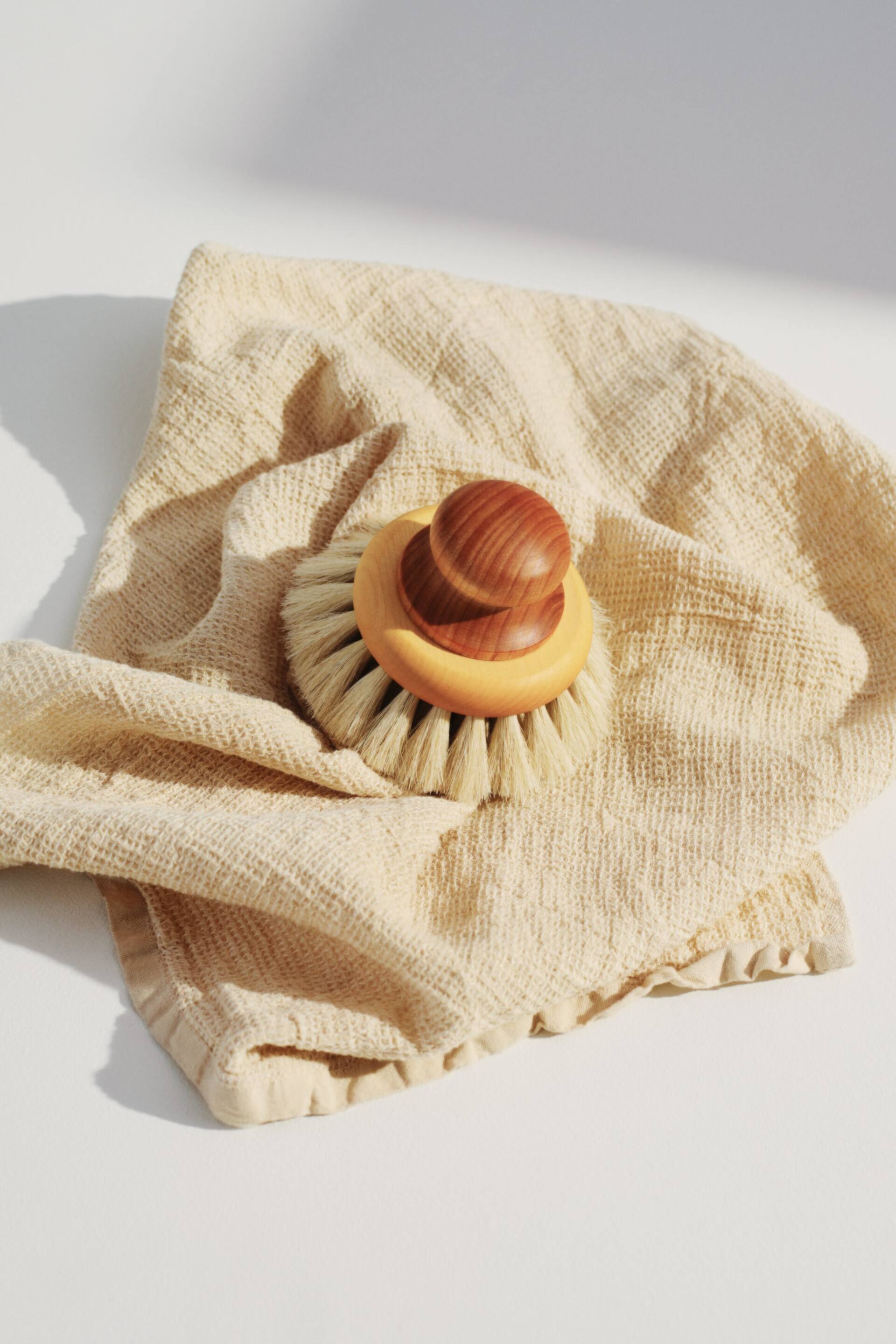 Agrandir l'image: Une brosse en bois repose sur un tissu texturé beige clair, présentée sous un léger angle de vue plongeante. La brosse arbore un manche arrondi brun foncé, une base en bois brun clair, et des poils de couleur naturelle qui rayonnent vers l'extérieur.