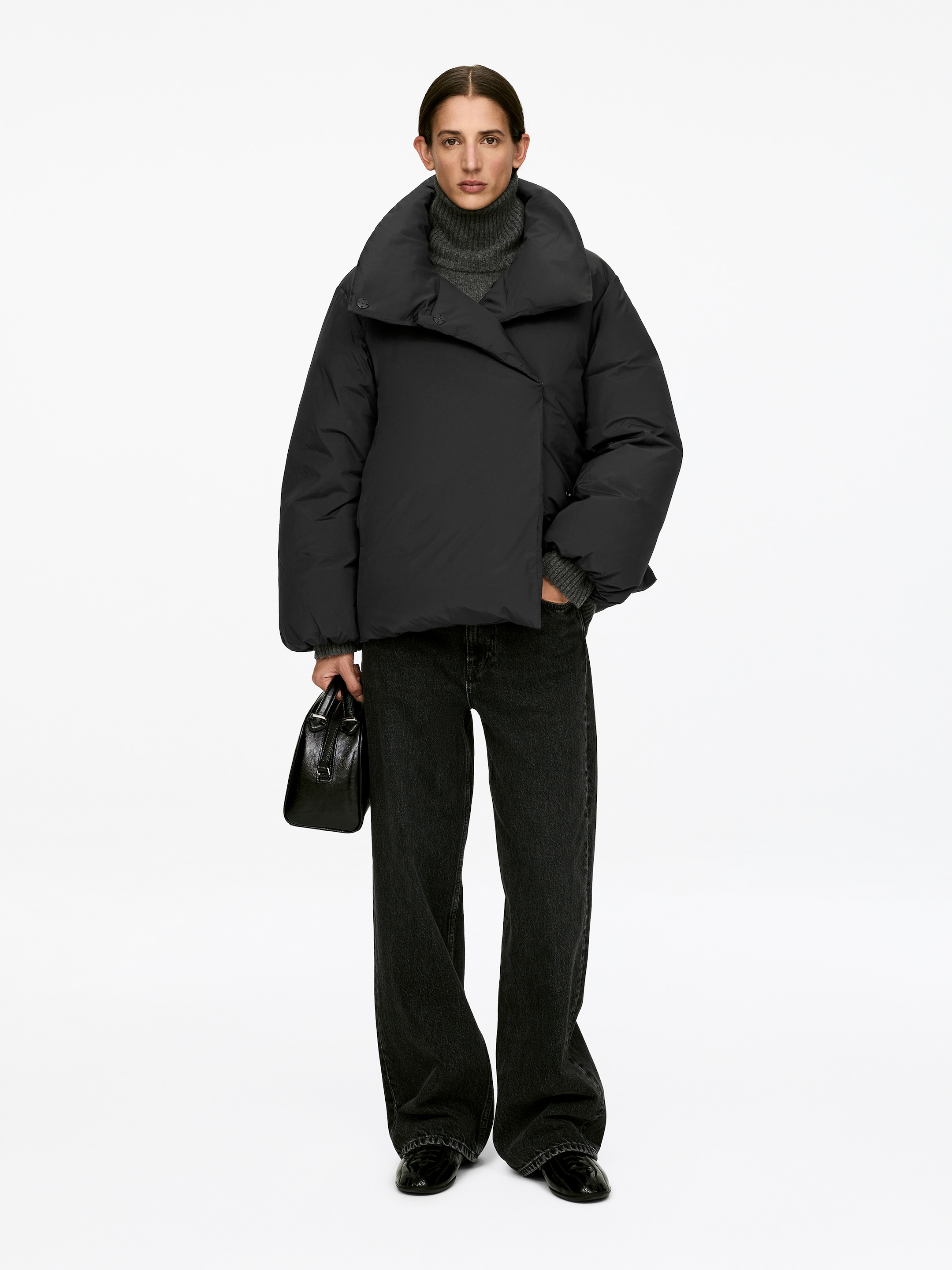 Agrandir l'image: Une veste matelassée noire, présentant une silhouette volumineuse et un col montant, est portée par le mannequin. Son devant est orné d’une fermeture asymétrique, superposée à un pull à col roulé gris à côtes visibles.