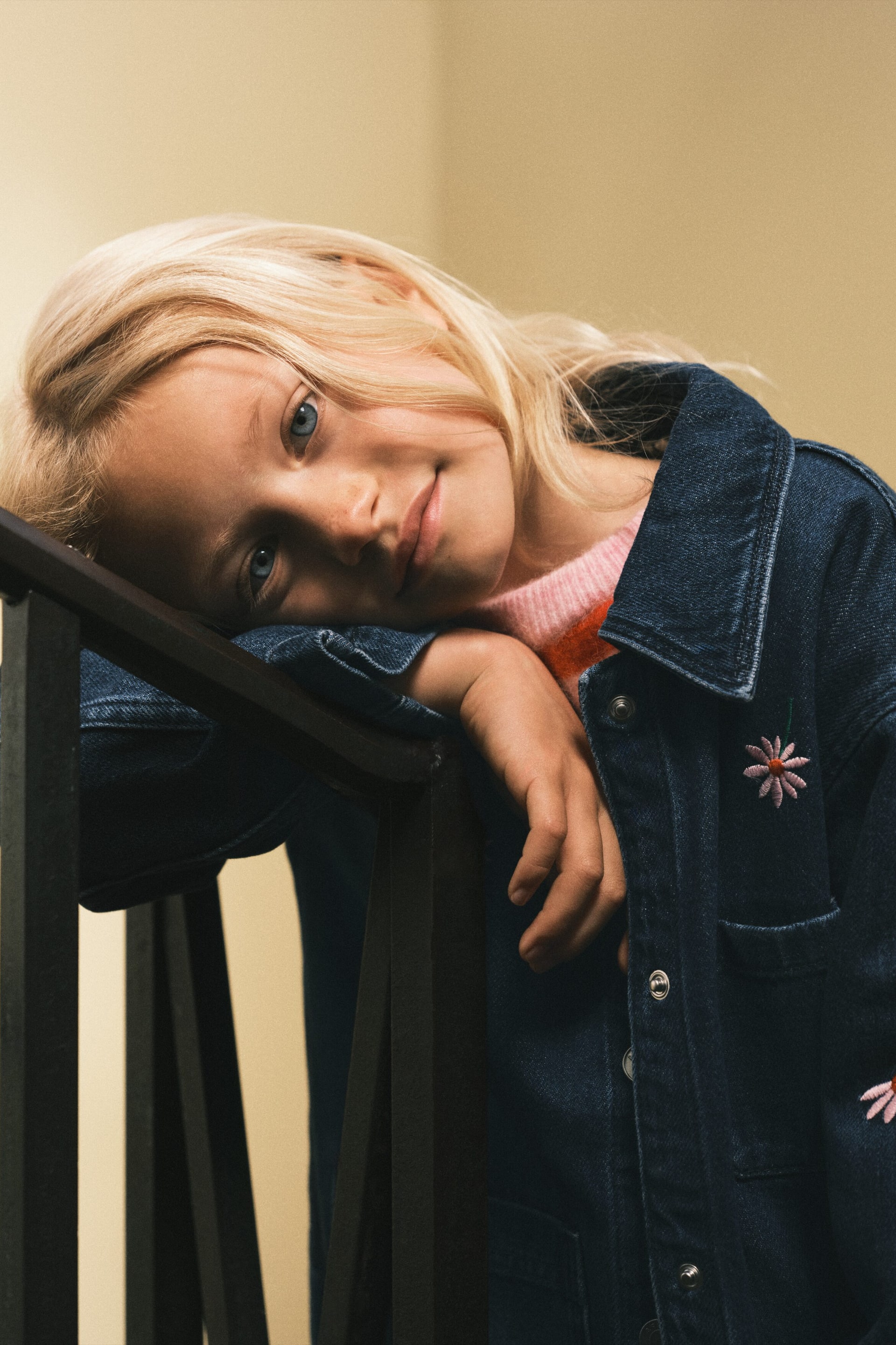 Se større billede: En mørkeblå denimjakke med spids krave, trykknaplukning foran og en brystlomme med klap og trykknap. Jakken er prydet med broderede tusindfryd i pink og hvid, og en pink- og orangestribet sweater kan ses under.