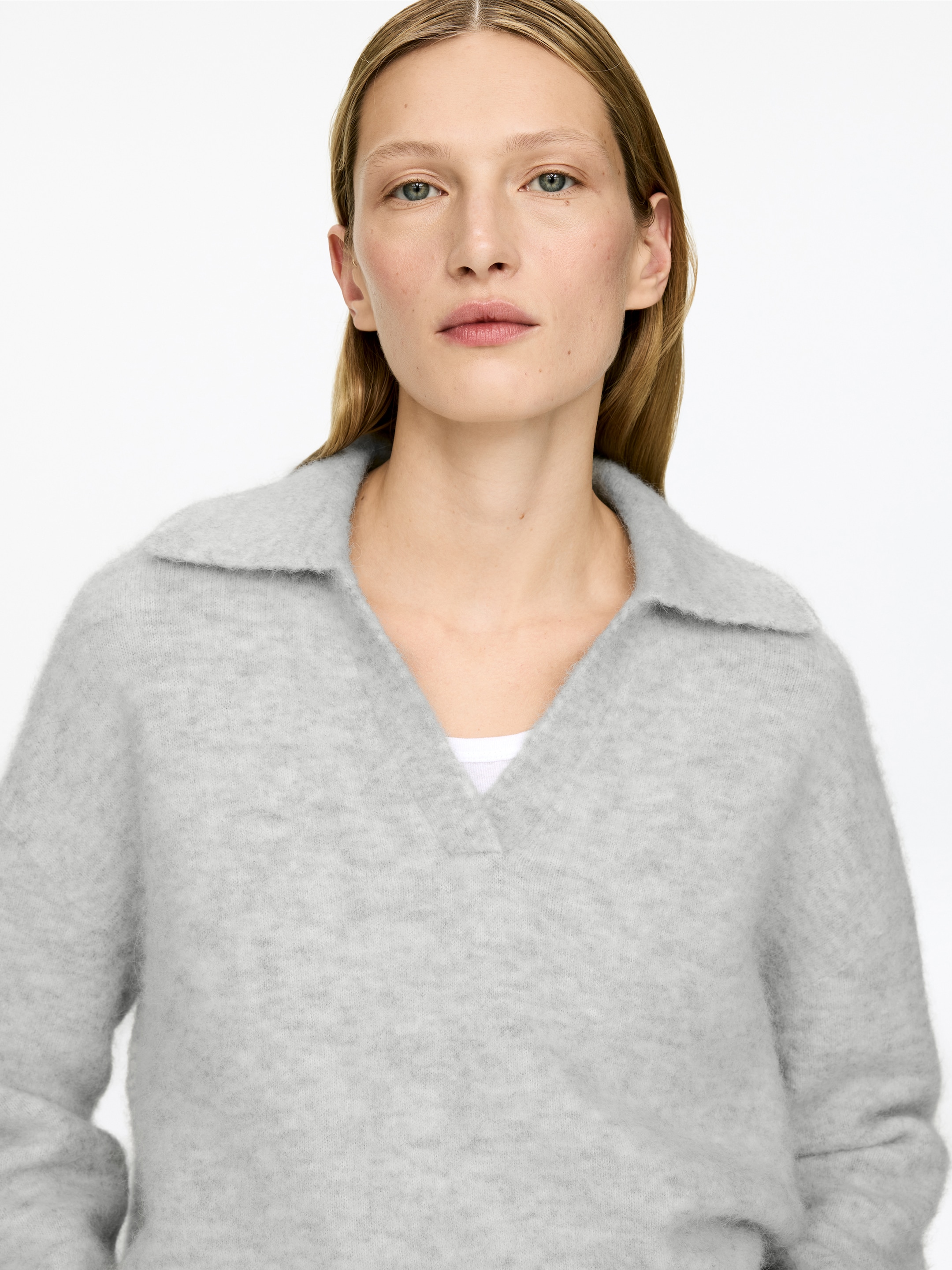 Agrandir l'image: Un pull en maille gris clair et duveteux présente une coupe décontractée, avec un col à revers et une ouverture en V, superposé sur un vêtement blanc.
