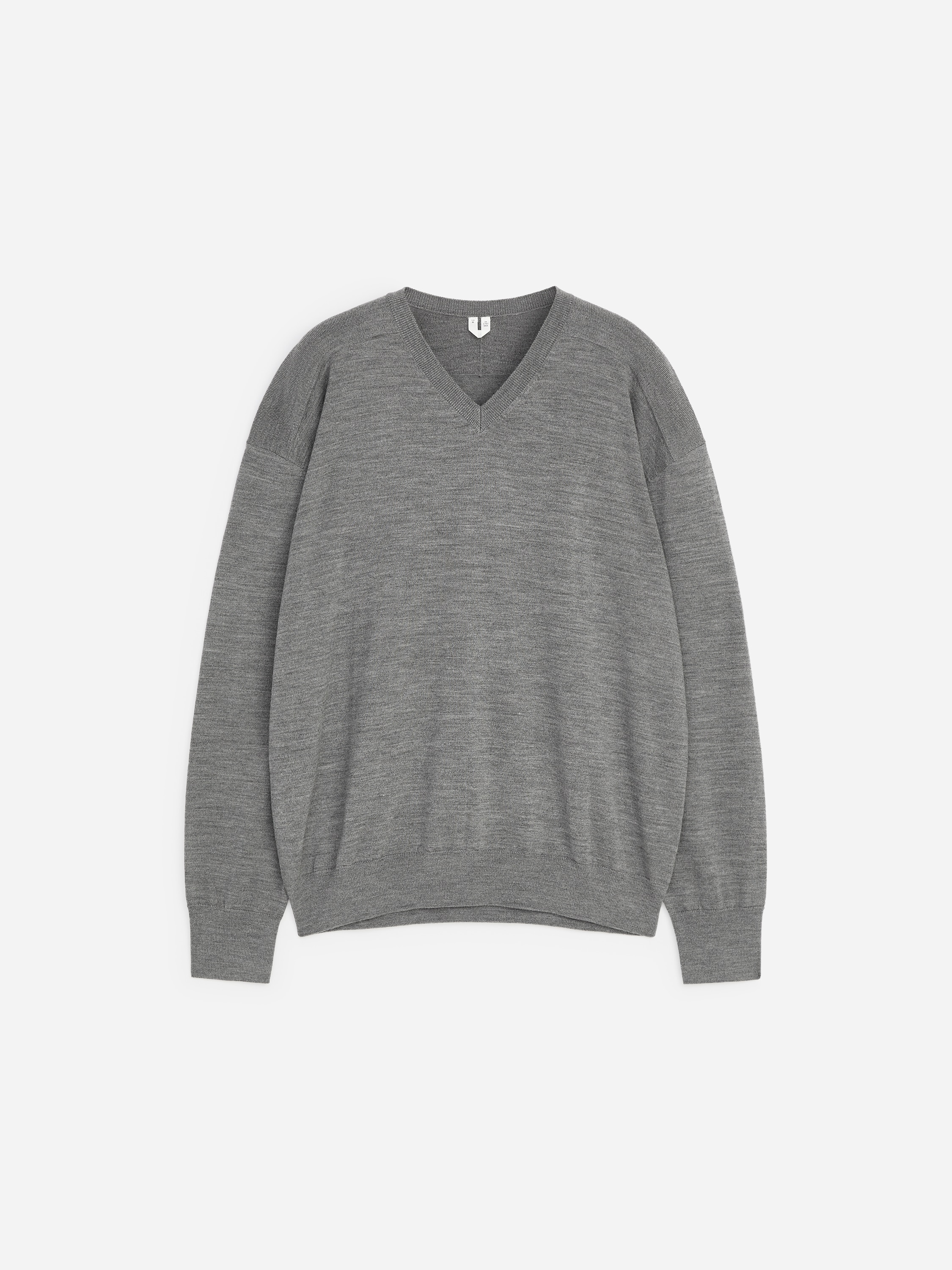 A grey, fine-knit V-neck jumper is being worn by the model, who is standing with their hands in the pockets of dark blue jeans. The jumper features a relaxed fit with dropped shoulders and ribbed detailing on the neckline, cuffs and hem.