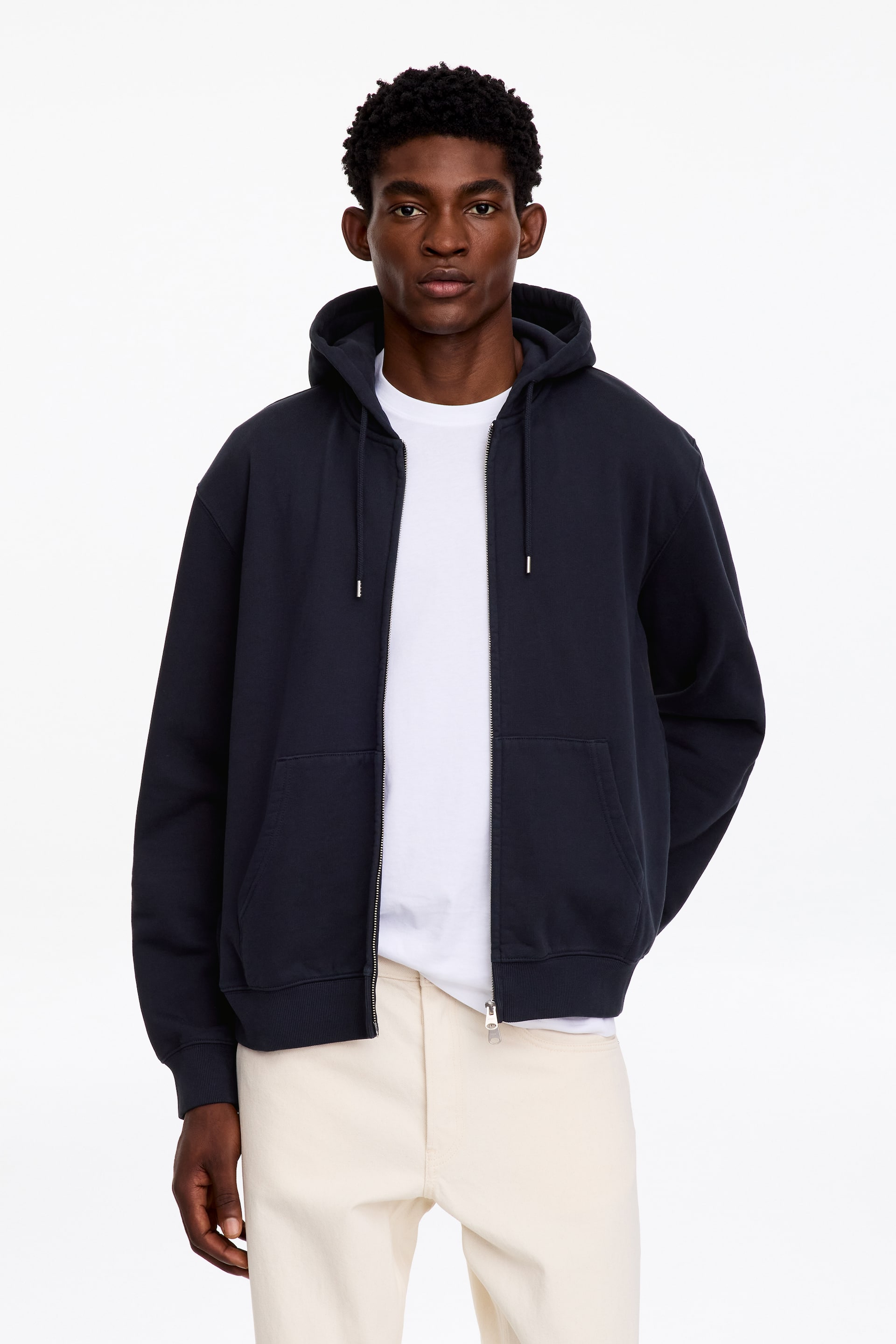 Ver imagem maior: O modelo está a usar um hoodie com fecho de correr em azul-marinho escuro por cima de uma t-shirt branca e calças em tom creme. O hoodie apresenta um fecho frontal bidirecional aberto, capuz com cordão, bolsos canguru divididos e acabamentos canelados nos punhos e na bainha.