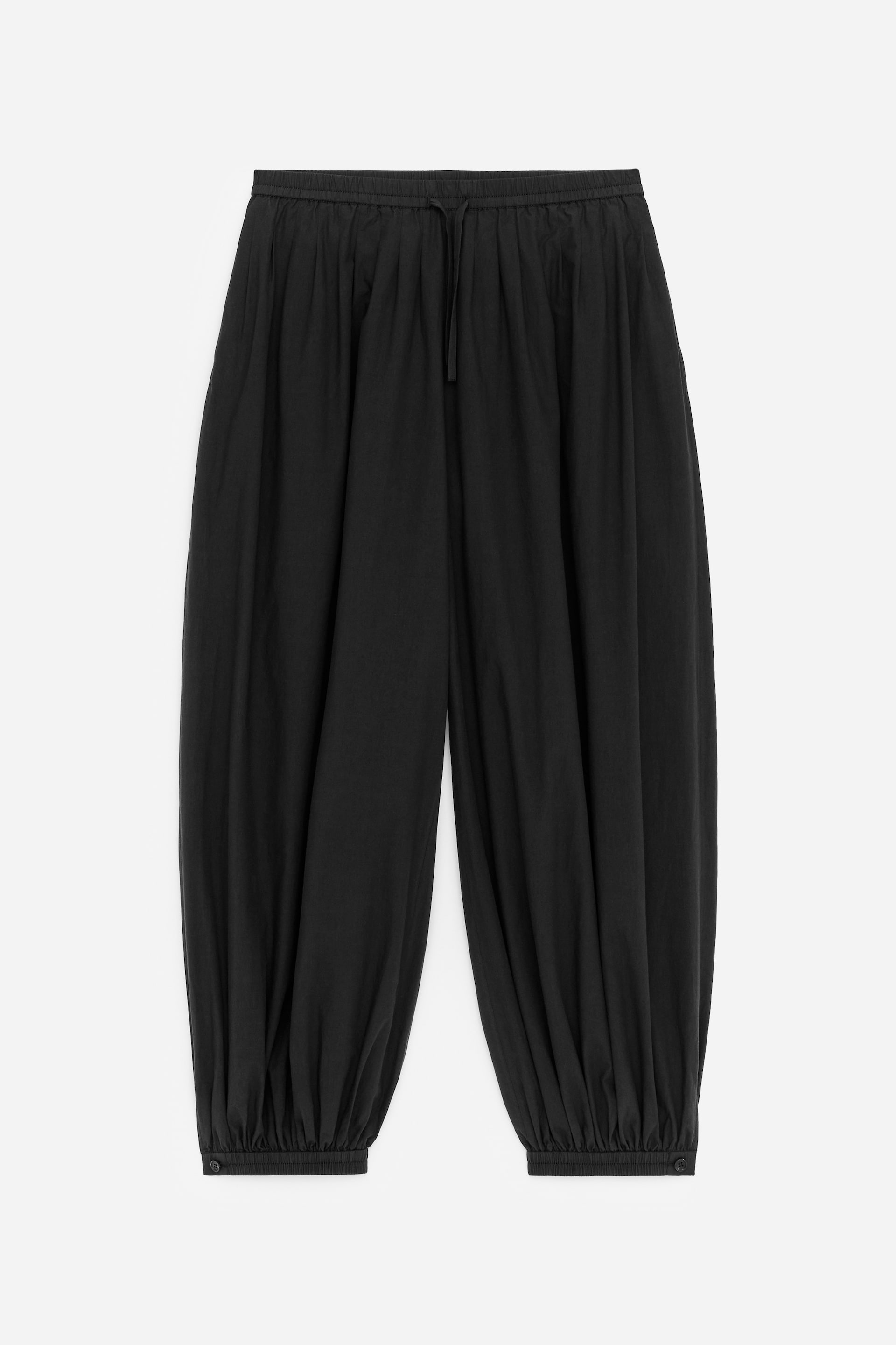 Visualizza immagine più grande: FRONTE: Pantaloni silhouette a palloncino neri, realizzati in popeline di cotone croccante, caratterizzati da una fascia elastica in vita con un delicato laccetto e morbide pieghe che si trasformano in plissettature intorno alla vita. I pantaloni presentano anche tasche laterali cucite e polsini alla caviglia arricciati, ciascuno dettagliato con un piccolo bottone.