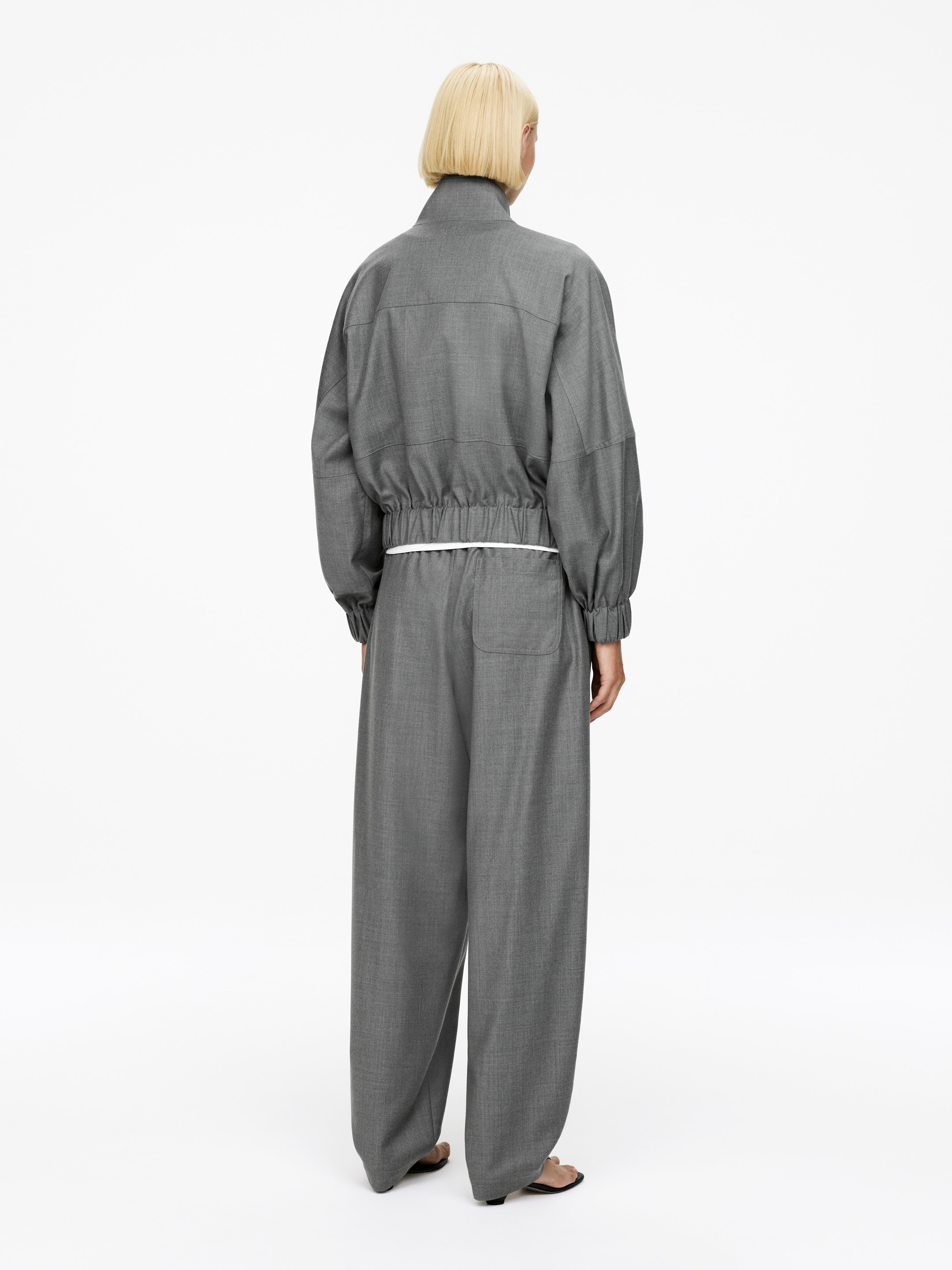 Grotere afbeelding bekijken: Een grijze jumpsuit wordt van de achterkant gepresenteerd, met een hoge opstaande kraag en lange mouwen met geplooide elastische manchetten. Het bovenste gedeelte is aangesnoerd bij een elastische taille met een wit trekkoord, waardoor een blouson-effect ontstaat over de wijde broekspijpen.