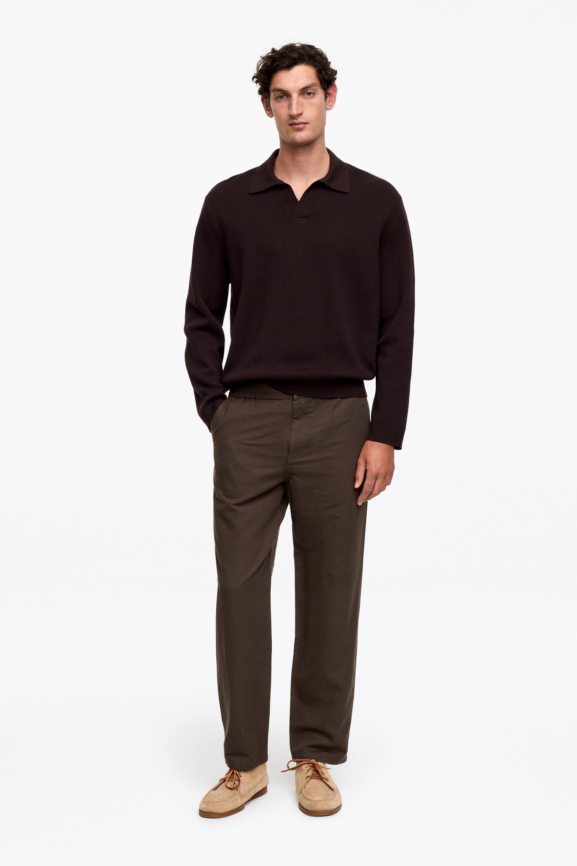 Vis større bilde: En mørkebrun strikket polosweater med spredt krage, V-hals plakkett og ribbestrikkede mansjetter og kant. Den presenteres fra en frontvendt vinkel sammen med olivengrønne bukser med avslappet passform, som viser en elastisk linning og rette ben, samt lysebrune mokkasin-stil sko i semsket skinn med synlig snøring.