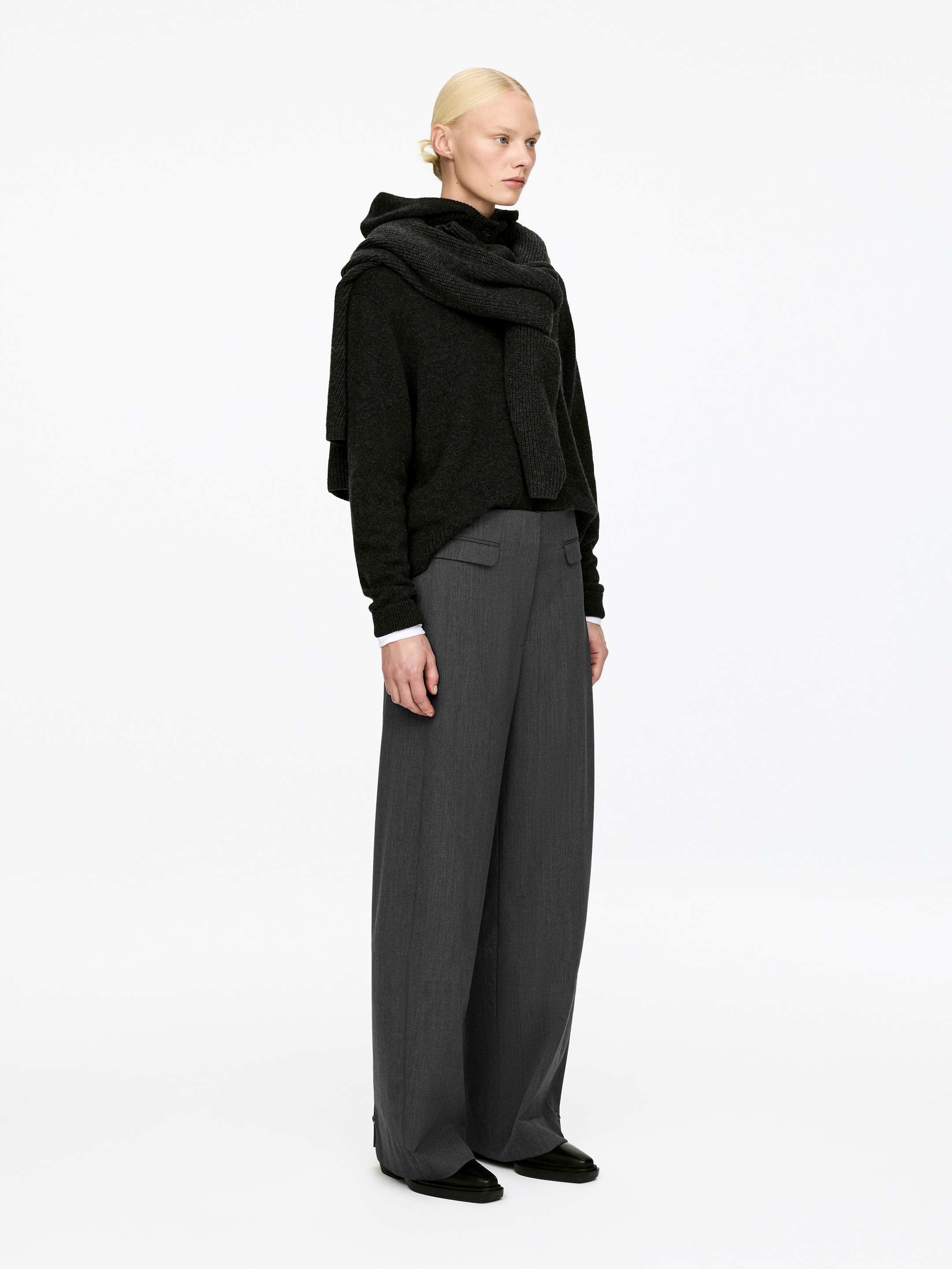 Visualizza immagine più grande: Un maglione a collo alto in maglia grigio scuro e una sciarpa abbinata sono indossati sopra i polsini di una camicia bianca e pantaloni ampi grigio scuro, presentati in vista laterale, con i pantaloni che mostrano pieghe frontali e tasche laterali.