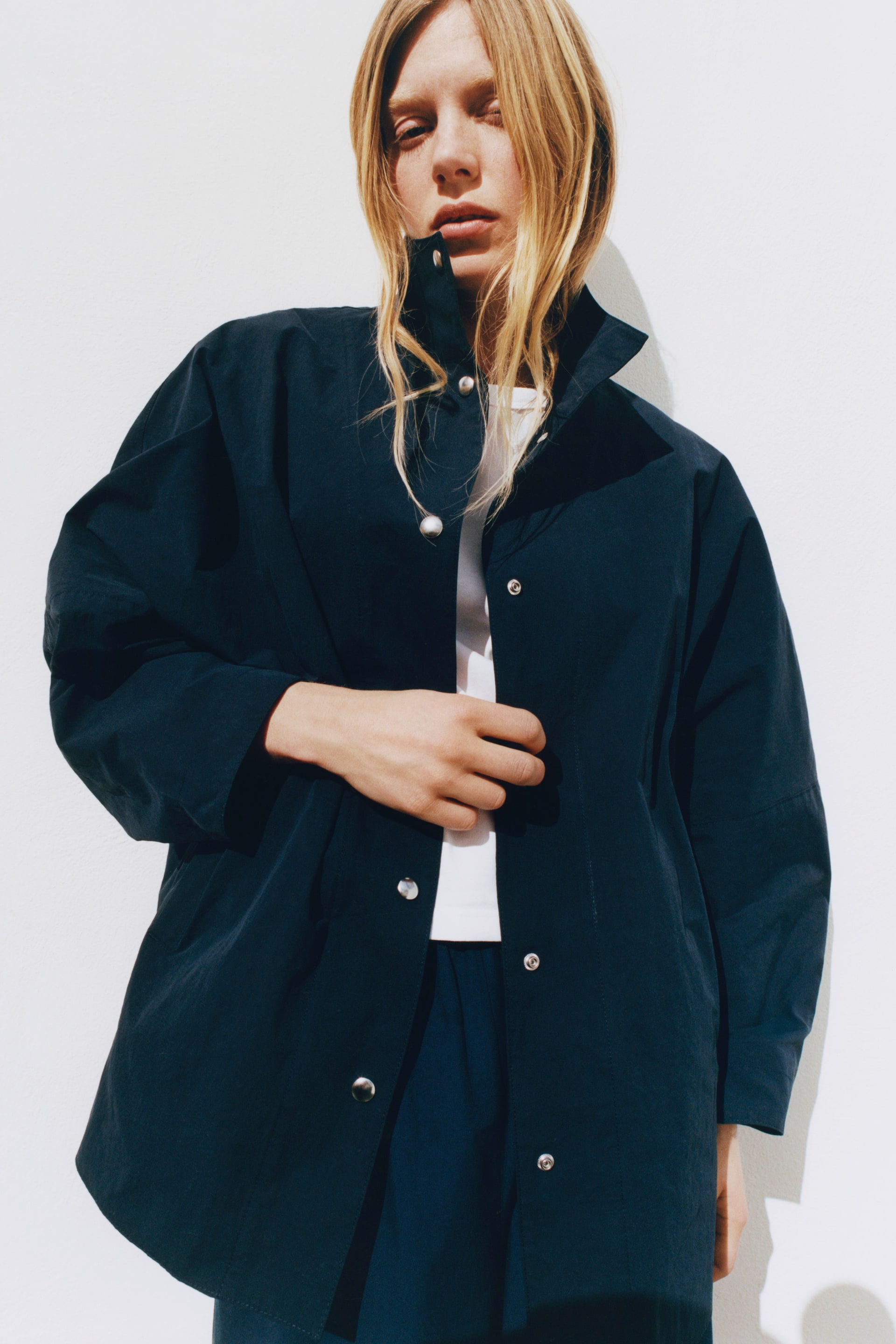 Grösseres Bild ansehen: Die dunkelblaue Jacke wird aus einer frontalen Perspektive präsentiert, leicht zur rechten Seite des Trägers geneigt, mit einem hochgestellten Stehkragen. Silberfarbene Druckknöpfe verlaufen entlang der vorderen Knopfleiste, und die Jacke wird über einem weißen T-Shirt und dunkelblauen Hosen getragen.