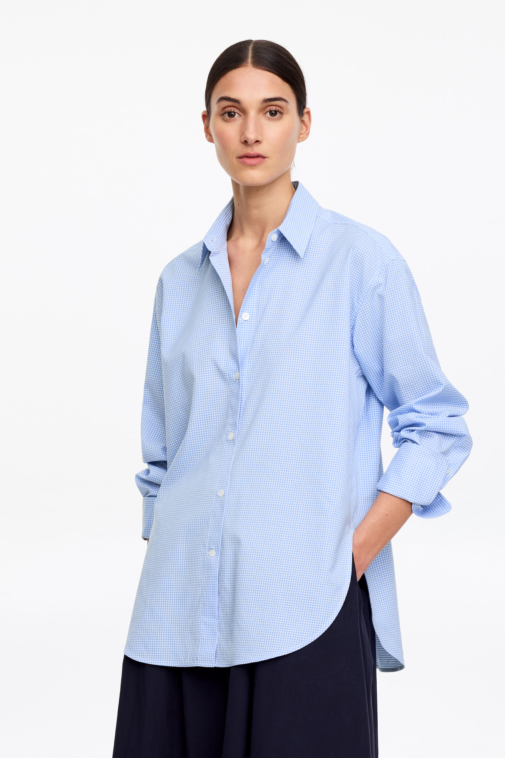 Grösseres Bild ansehen: Ein langärmeliges Hemd aus knackigem Baumwollpopeline mit einem hellblauen Vichy-Karo-Muster wird vom Model getragen. Dieser Button-up-Stil, einschließlich eines Haifischkragens, geknöpfter Ärmelbündchen und geöffneten oberen Knöpfen, präsentiert eine entspannte, aber dennoch elegante Silhouette. Das Model steckt eine Hand in die Tasche einer weit geschnittenen dunkelblauen Hose, wobei der abgerundete Saum des Hemdes sichtbar ungesäumt bleibt.