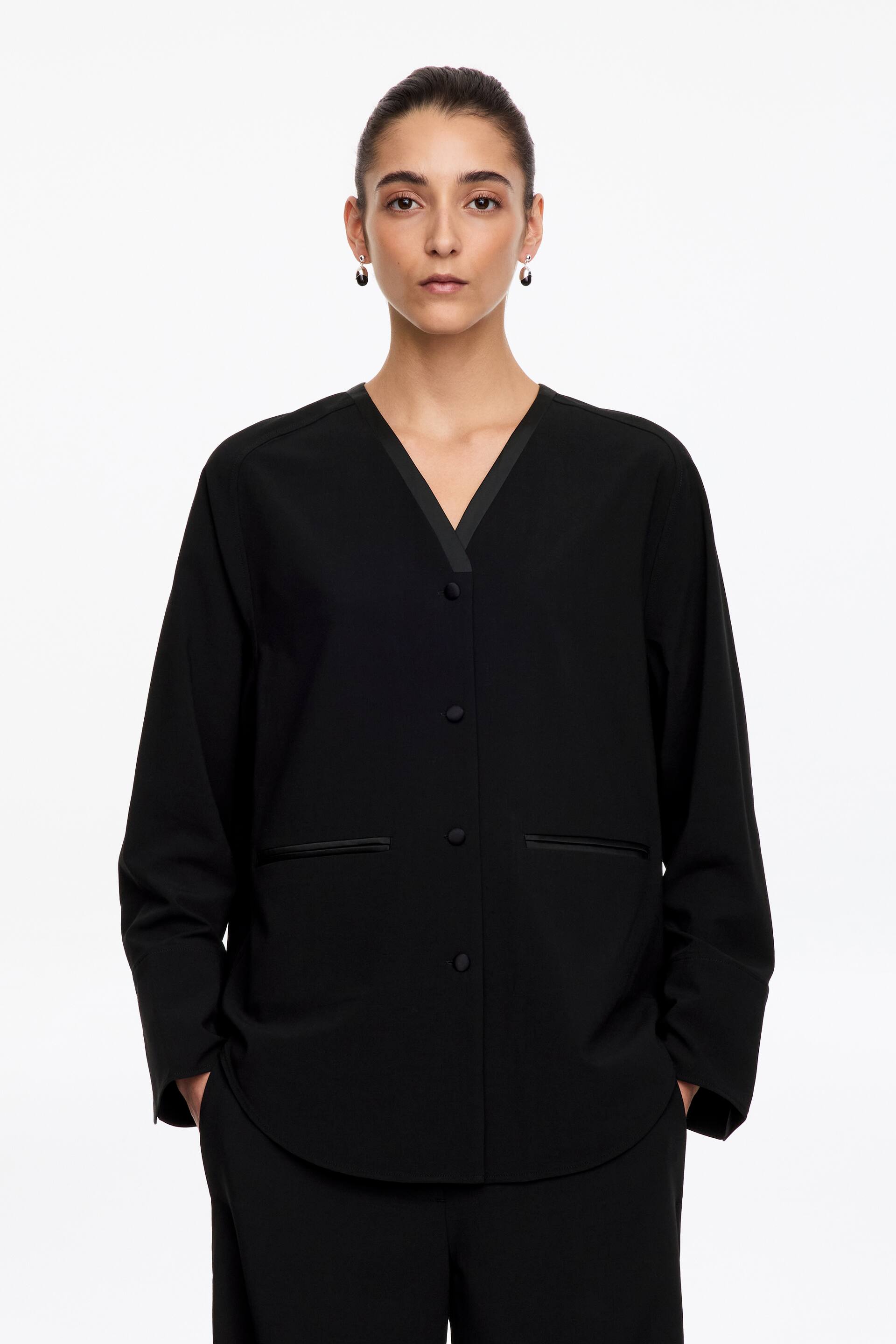 Grotere afbeelding bekijken: Een zwart tuxedo-overhemdblazer met relaxed fit, voorzien van een met satijn afgewerkte V-halslijn, met stof beklede knopen aan de voorkant en dubbele paspelzakken, wordt gedragen door het model. Het model staat recht vooruit met de handen in de zakken van een bijpassende zwarte pantalon.