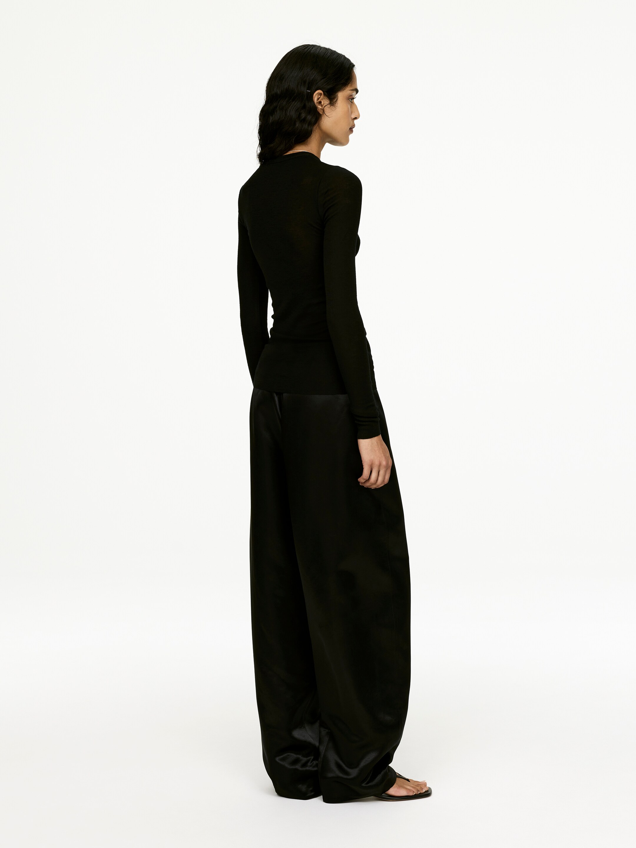 View larger image: The black, form-fitting, long-sleeved top with a high neckline is being presented from a side angle, styled with lustrous black wide-leg pants that are pooling at the ankles.