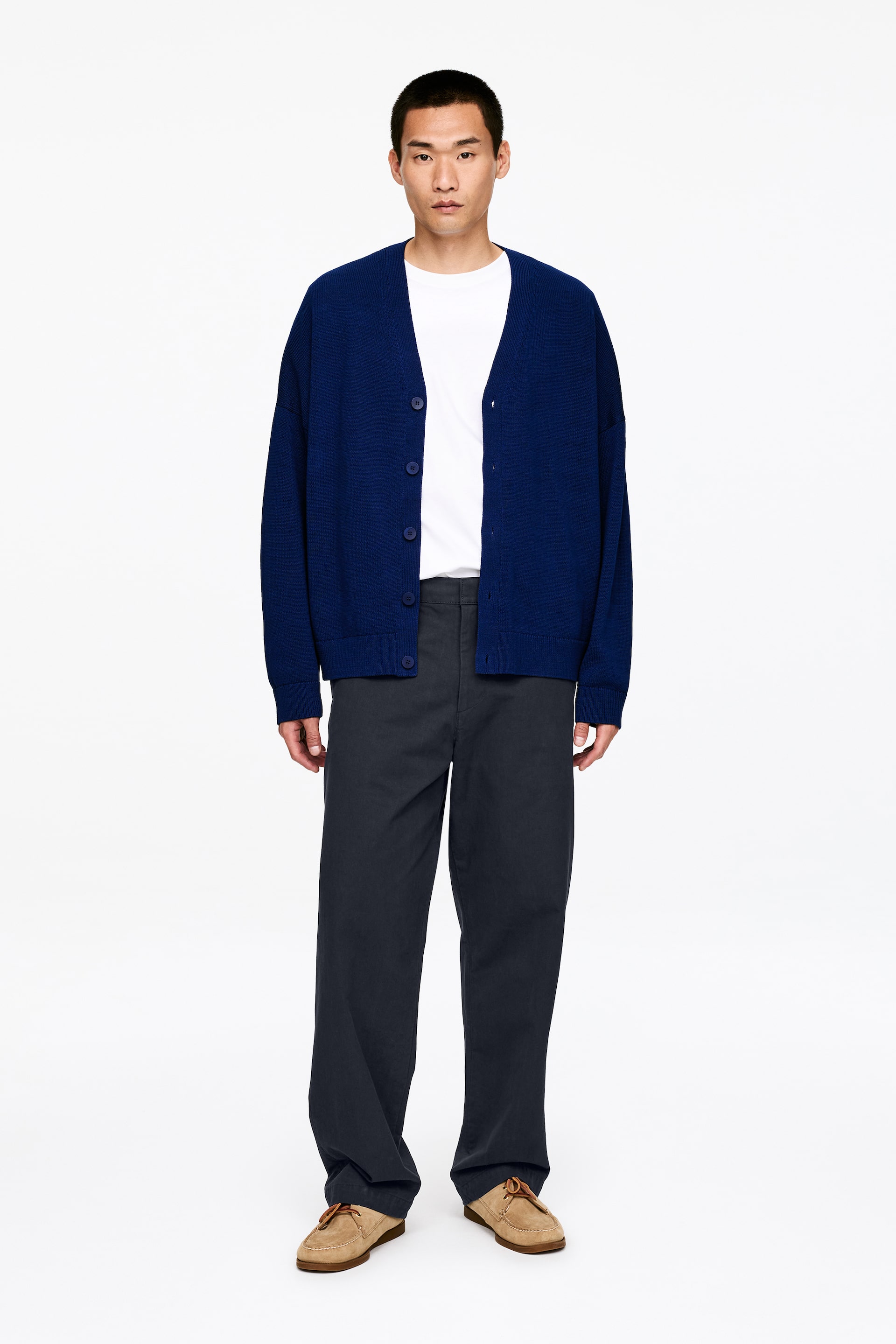 Vezi imaginea mai mare: Un cardigan albastru regal cu decolteu în V este prezentat din față, purtat deschis peste un tricou alb cu decolteu rotund. Acesta are nasturi asortați de culoare albastră, umeri căzuți și mâneci largi. Pantaloni gri închis cu croială dreaptă și mocasini din piele întoarsă de culoare cafeniu completează ținuta.