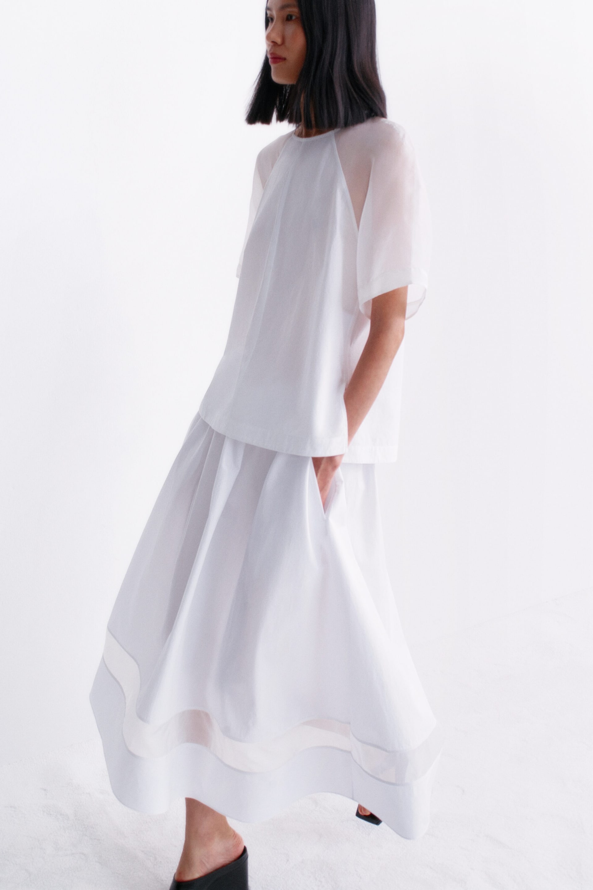 View larger image: A white two-piece outfit featuring a short-sleeved top with sheer raglan sleeves and a flowing midi skirt with sheer horizontal panels near the hem. The outfit is angled slightly to the left, showcasing its loose fit and layered design.