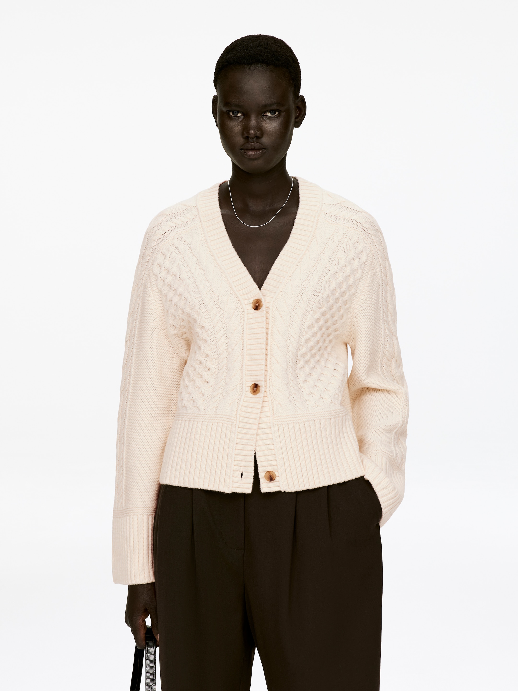 Agrandir l'image: Un cardigan en maille torsadée de couleur crème est porté par le mannequin, avec un col en V, une fermeture à trois boutons et des bordures côtelées. Deux des boutons du cardigan sont fermés, et il est porté par-dessus des vêtements sombres.