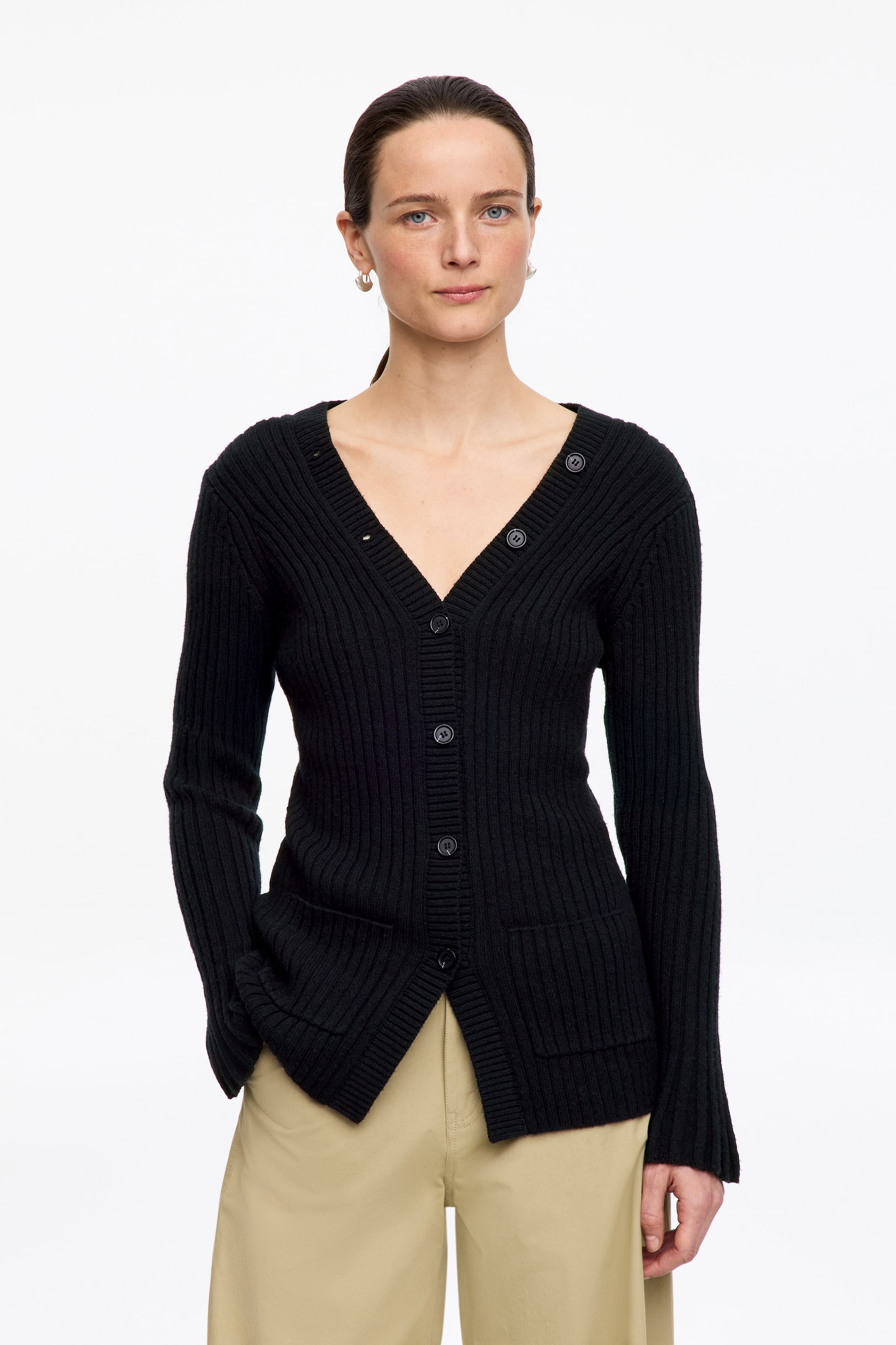 Visualizza immagine più grande: Un cardigan nero a maglia a coste è indossato, caratterizzato da uno scollo a V, una chiusura frontale con bottoni tondi scuri, maniche lunghe a coste e due tasche applicate sull'orlo anteriore.