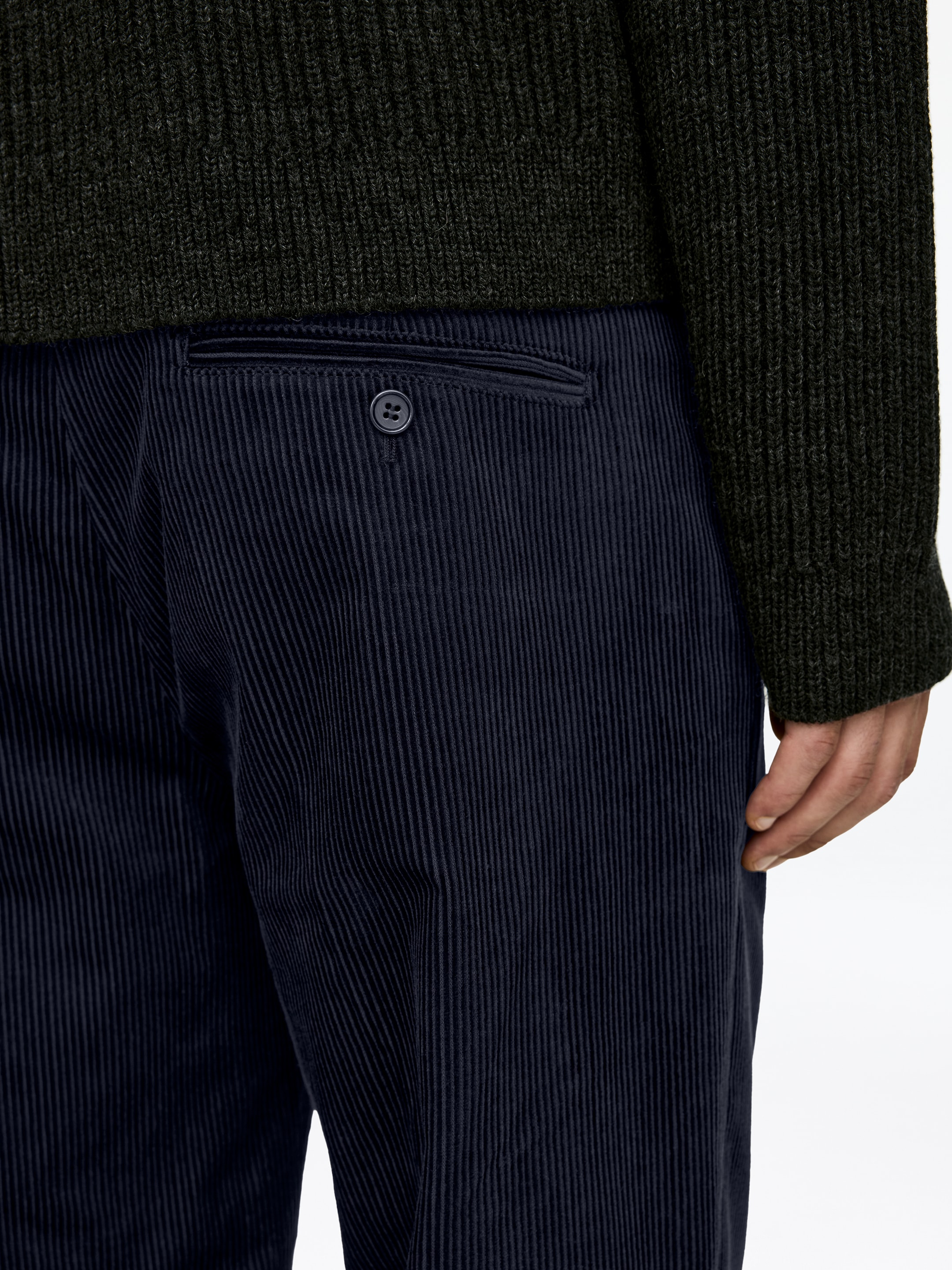 Ingrandisci l'immagine: I pantaloni in velluto a coste blu navy sono mostrati da un angolo laterale-posteriore, mettendo in evidenza le linee verticali e una tasca posteriore a filetto. La tasca è dettagliata con un singolo bottone scuro, mentre un maglione a maglia verde scuro è stratificato sopra la cintura dei pantaloni.