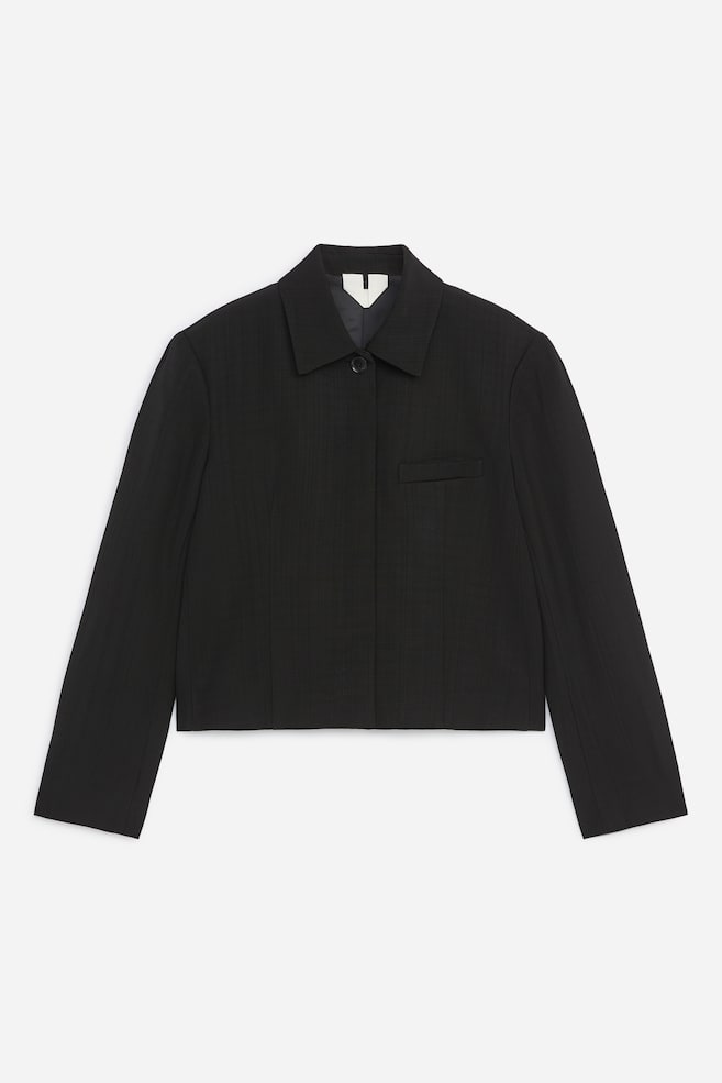 Eine schwarze, kurz geschnittene, taillierte Jacke ist von vorn zu sehen und präsentiert eine strukturierte Silhouette mit spitzem Reverskragen und einem einzelnen sichtbaren oberen Knopf über einer verdeckten Knopfleiste. Die Jacke verfügt über lange Ãrmel, dezente vertikale Abnäher zur Formgebung und eine einzelne horizontale Leistentasche auf der linken Brust. - 1