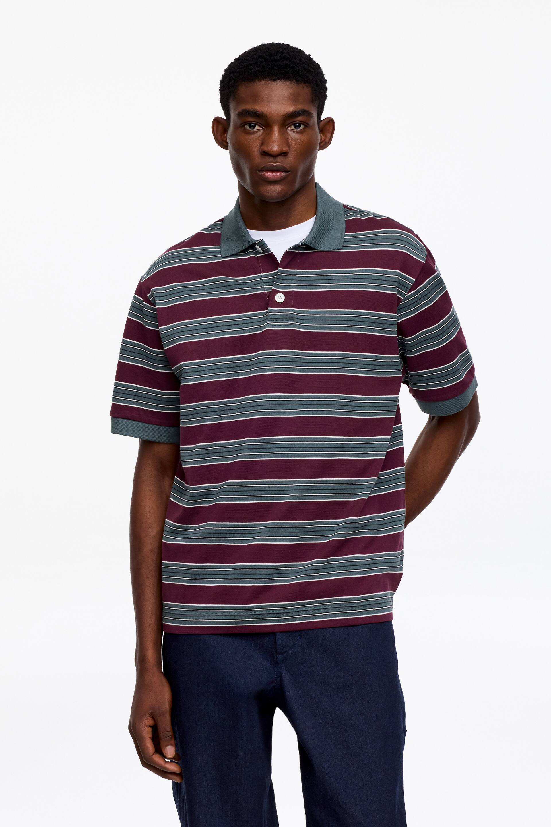 View larger image: A striped polo shirt is facing forward, showcasing wide horizontal stripes in burgundy, dark teal, and thin white. It features a solid dark teal collar and matching short sleeve cuffs, with a two-button placket visible at the front, worn over a white undershirt.