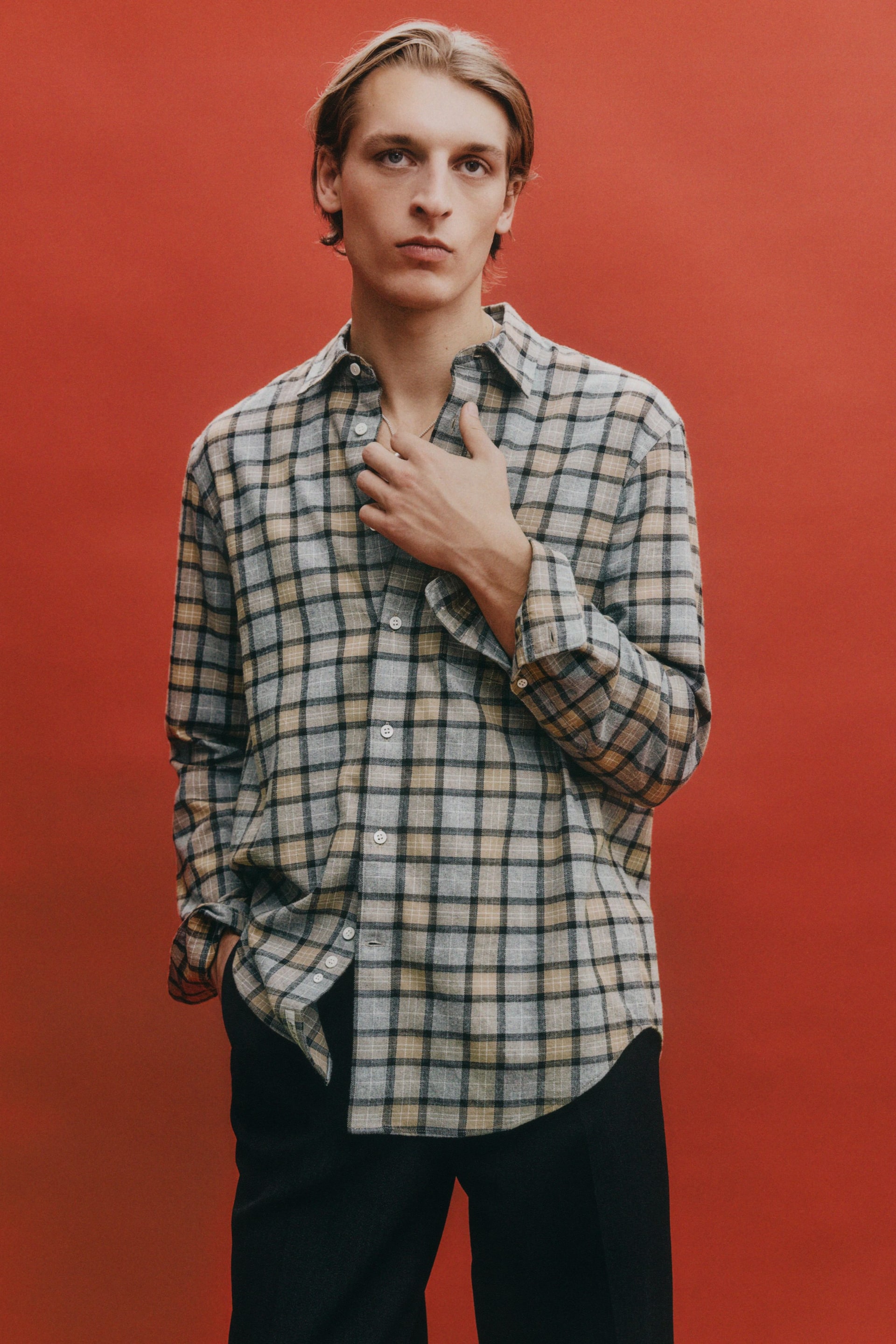 Zobrazit větší obrázek: Přední strana kostkované košile s šedým, černým a béžovým kostkovaným vzorem. Je navržena s špičatým límcem, dlouhými rukávy s manžetami na knoflíky a zakřiveným lemem. Bílé knoflíky vedou dolů po přední léze a košile má volný střih s mírně otevřeným límcem.