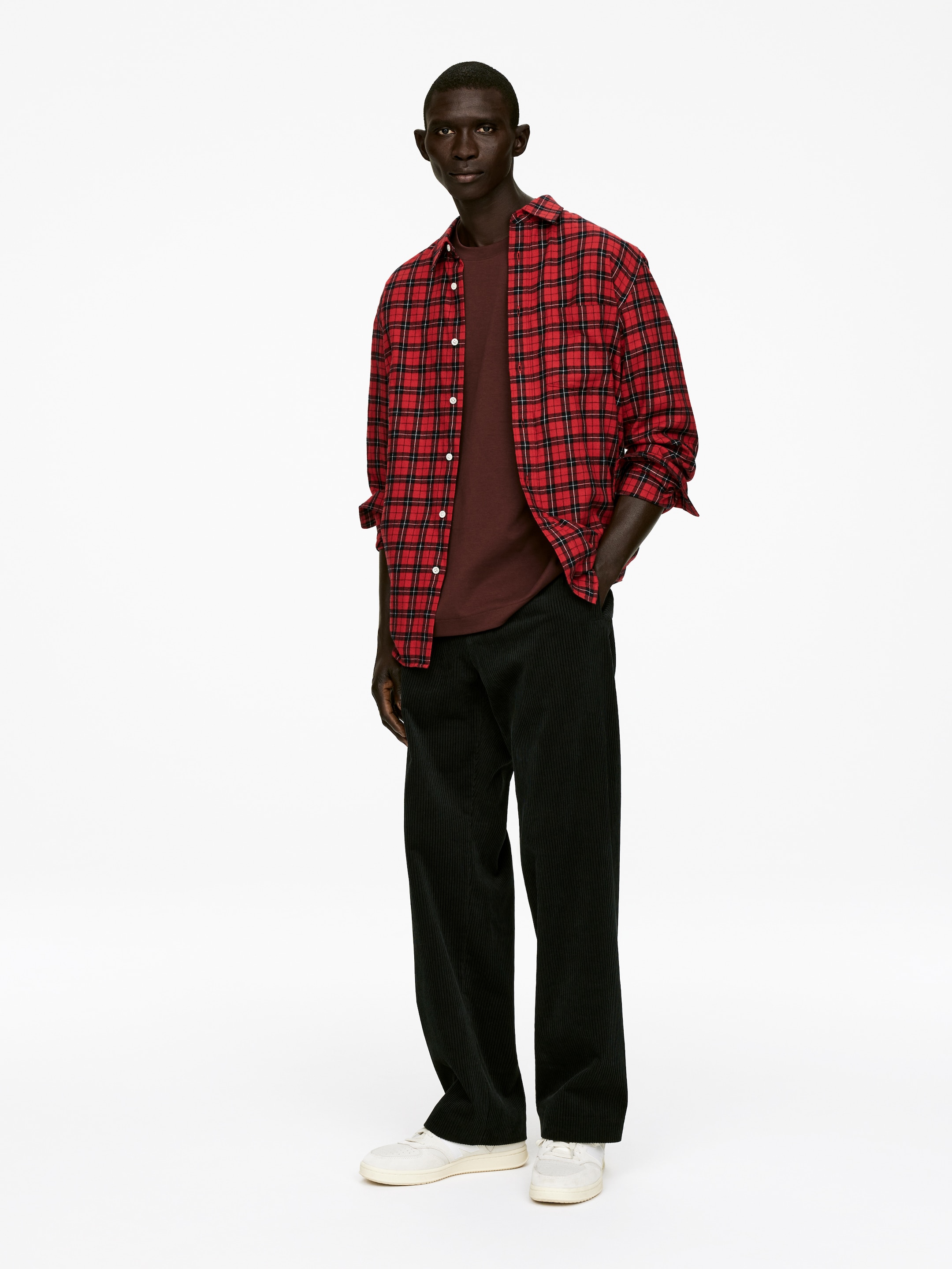 Visa större bild: En outfit presenteras från en något vinklad framifrån vy, med en öppen röd och svart rutig skjorta med krage och upprullade ärmar som bärs över en solid mörkbrun t-shirt med rund hals. Skjortan och t-shirten kombineras med mörkgröna breda manchesterbyxor och vita låga sneakers.