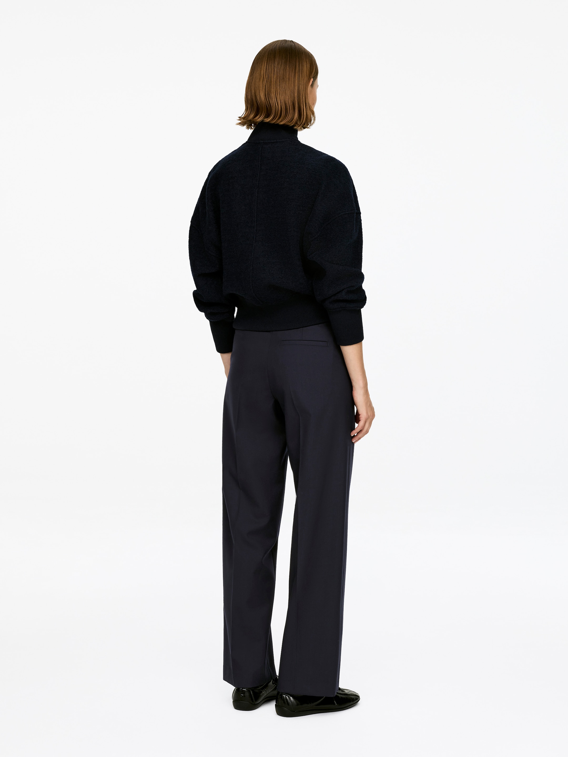 Agrandir l'image: Un pantalon en laine bleu marine foncé, avec un tissage hopsack, une coupe mi-haute et des plis marqués, est porté par le mannequin. Ils sont stylisés avec un haut foncé et des chaussures plates noires.