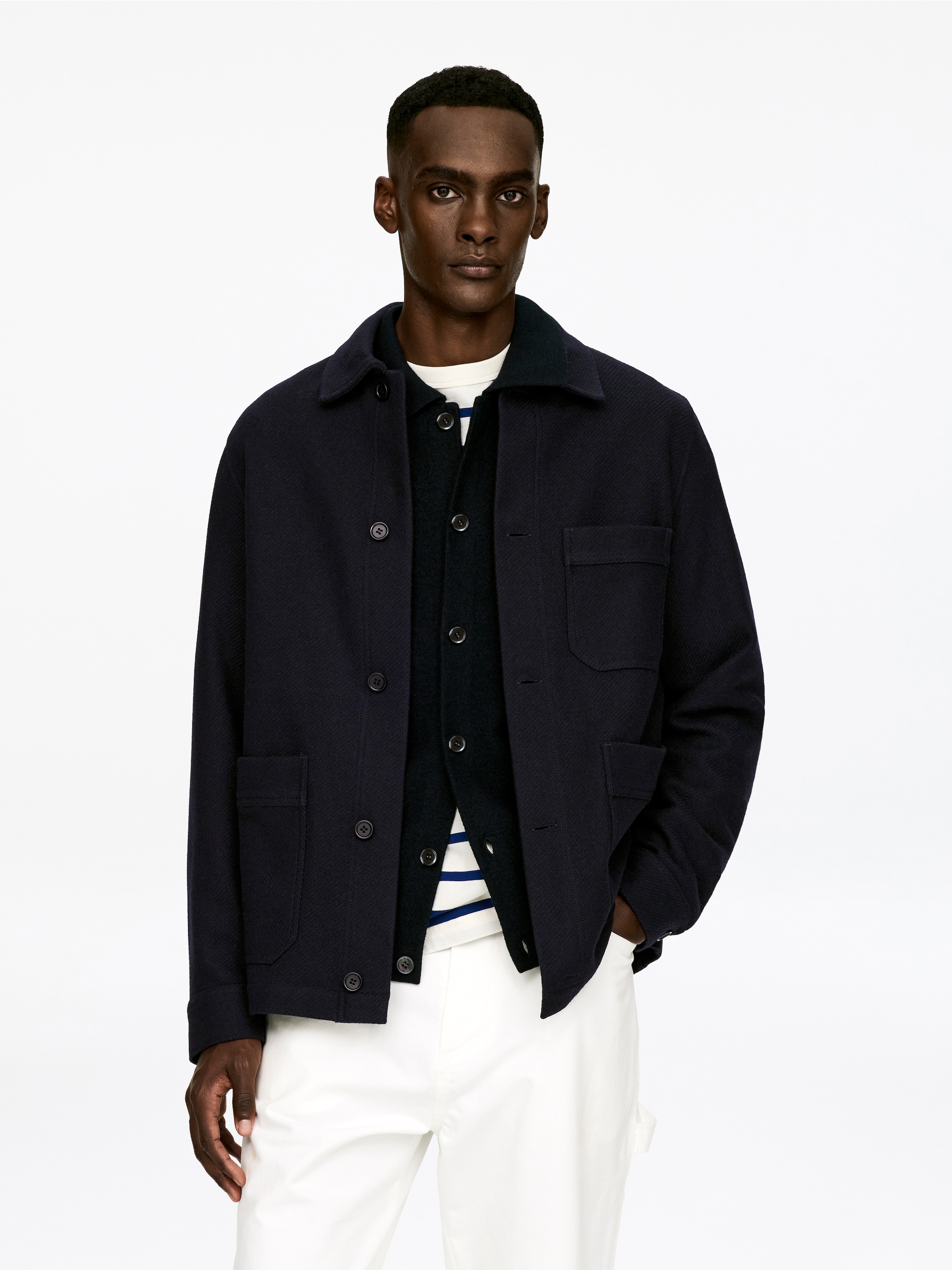 Ver imagen más grande: Una chaqueta estilo sobrecamisa de color azul marino está orientada hacia adelante, con un clásico cuello de camisa, un cierre frontal con botones oscuros, un bolsillo de parche en el pecho y dos bolsillos de parche más grandes en la parte inferior. Está superpuesta a un cárdigan oscuro con cierre de botones y una camisa de manga larga crema con rayas horizontales.