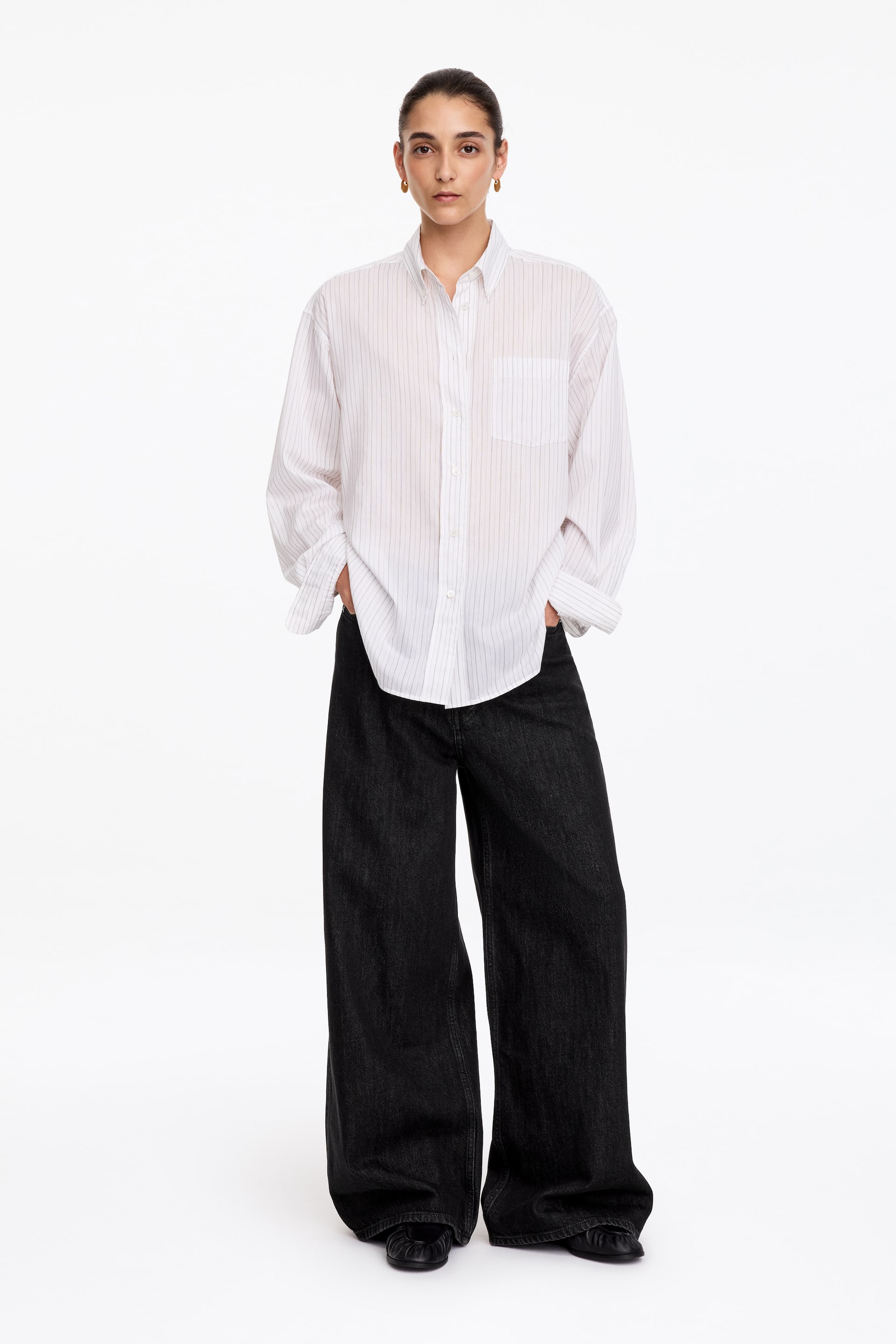 Agrandir l'image: Une chemise blanche à manches longues et boutonnée, à fines rayures verticales, est portée par le mannequin, stylée rentrée à l’extérieur sur un jean noir à jambes larges. La chemise présente un col classique et une poche plaquée sur la poitrine, tandis que les mains du mannequin reposent dans les poches du jean.