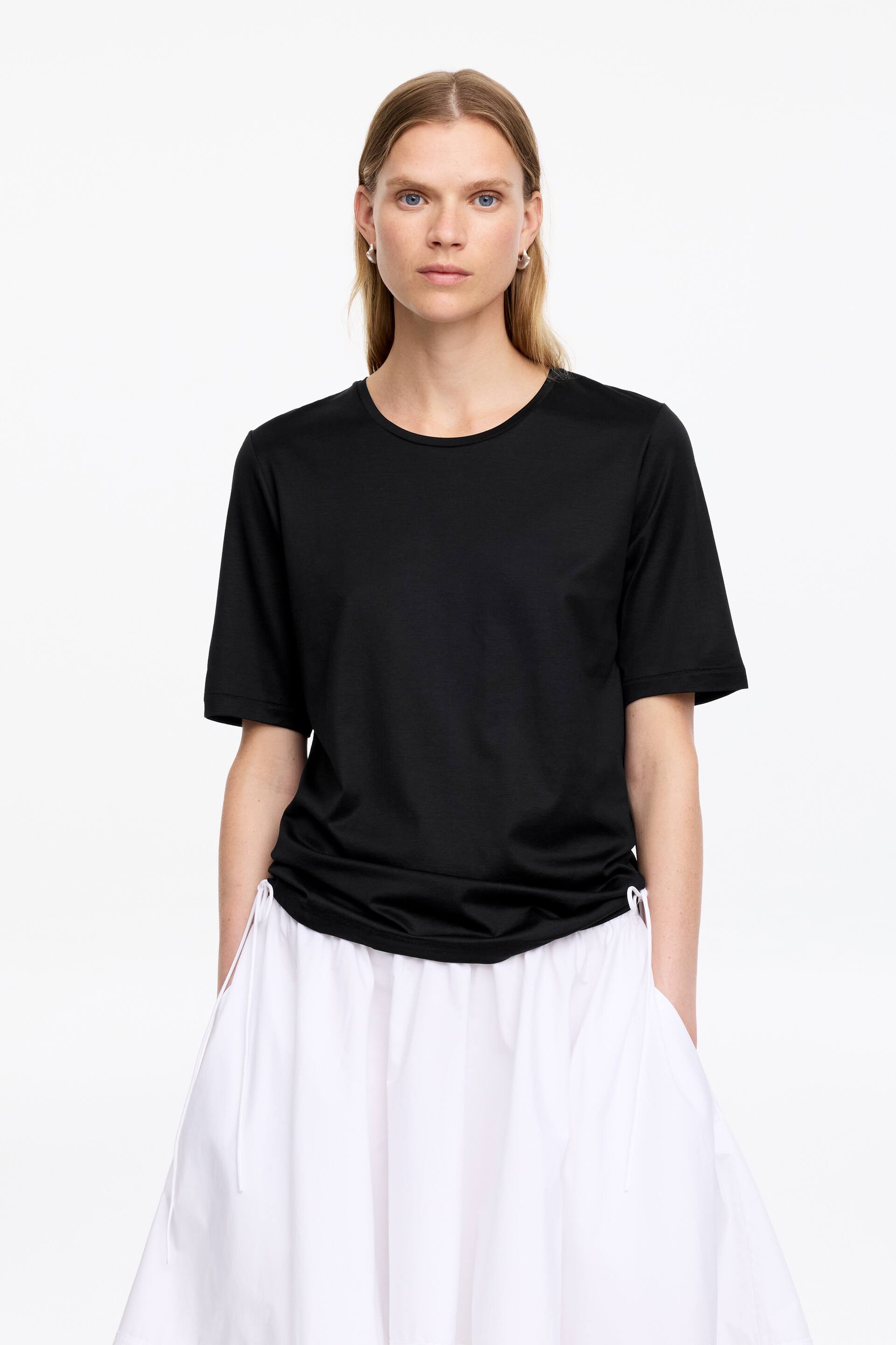 Visualizza l'immagine ingrandita: Una t-shirt nera è rivolta in avanti, caratterizzata da un girocollo e maniche corte. Il suo orlo rilassato è raccolto ai lati, creando un effetto arricciato sulla gonna bianca a strati.