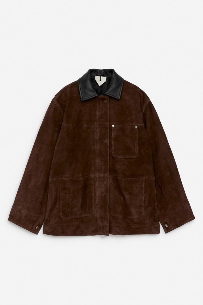 The FRONT of an oversized brown suede jacket is featuring a contrasting black collar, two large patch pockets with flaps on the lower front, and a single patch pocket on the upper left chest. A central placket is concealing a zip closure and hidden press buttons, with one visible button near the collar, while tabs with silver-colored shank buttons are visible at the side hems for fit adjustments. - 1