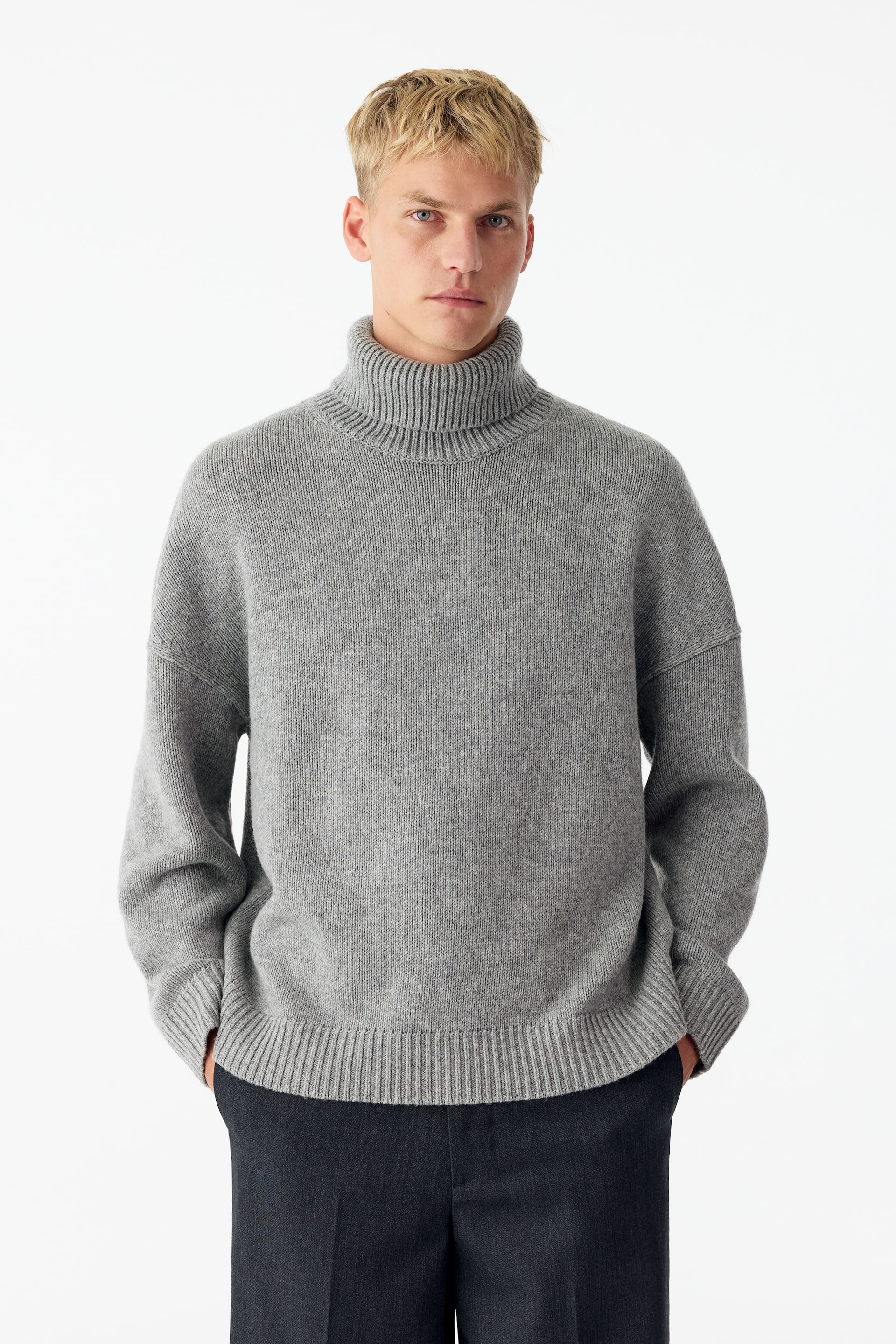 View larger image: A grey roll-neck wool jumper featuring a relaxed fit, dropped shoulders, and ribbed trims is being worn by the model. The model is standing with hands placed in the pockets of dark trousers.